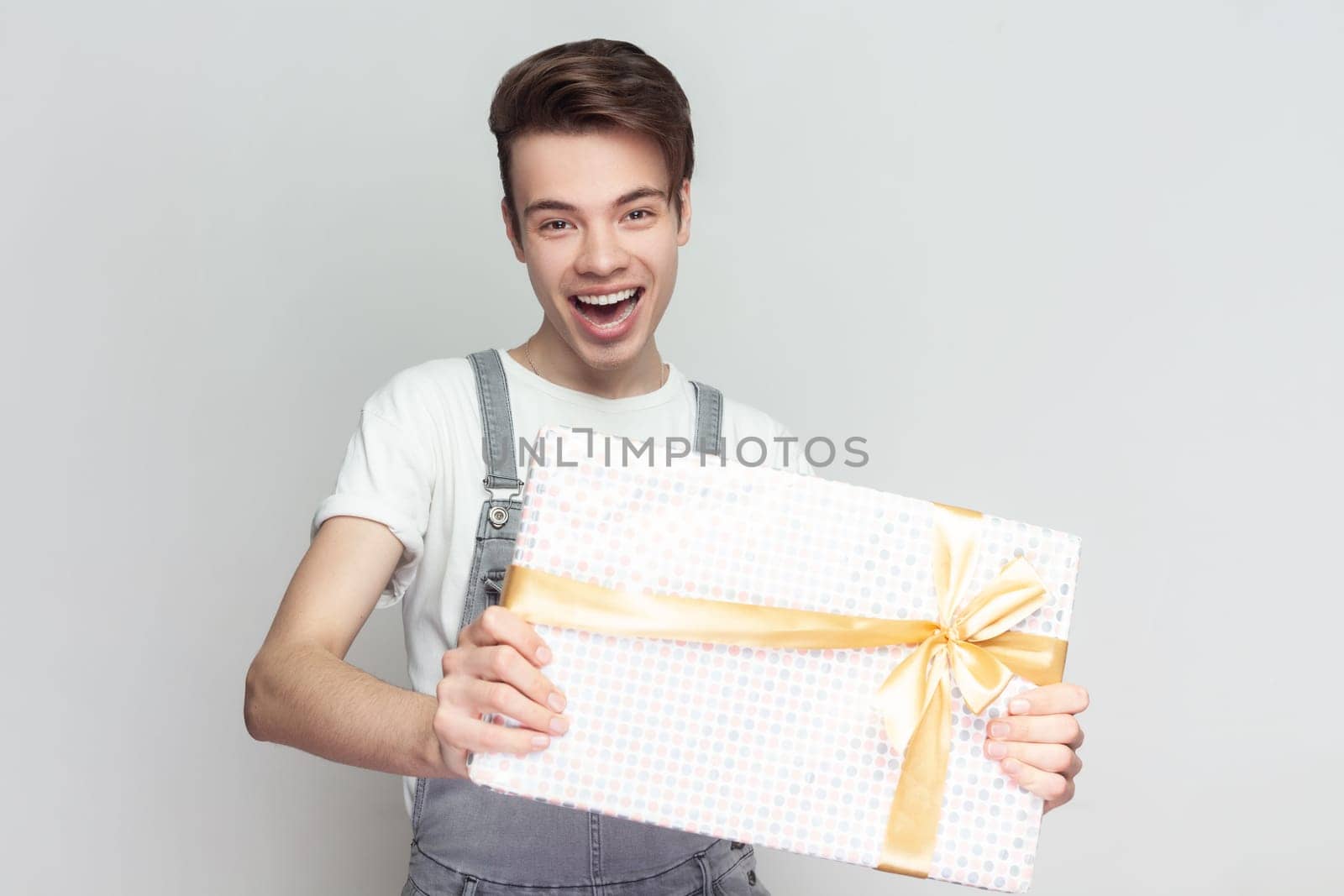 Portrait of joyful attractive excited brunette man standing standing with present box in hands, congratulating with holiday, wearing denim overalls. Indoor studio shot isolated on gray background.