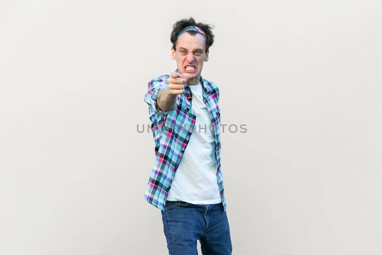 Furious angry man looses temper, screams and points at you, blames someone and expresses negative emotions, wearing checkered shirt and headband. Indoor studio shot isolated on gray background.