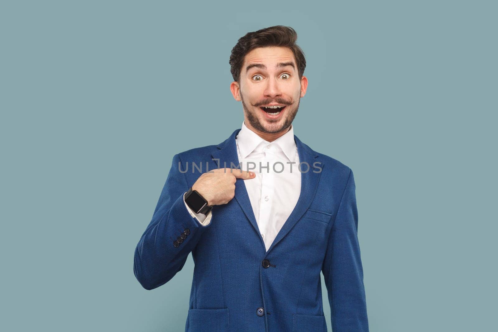 Portrait of excited amazed handsome man with mustache standing pointing at himself with surprised face, wearing official style suit. Indoor studio shot isolated on light blue background.