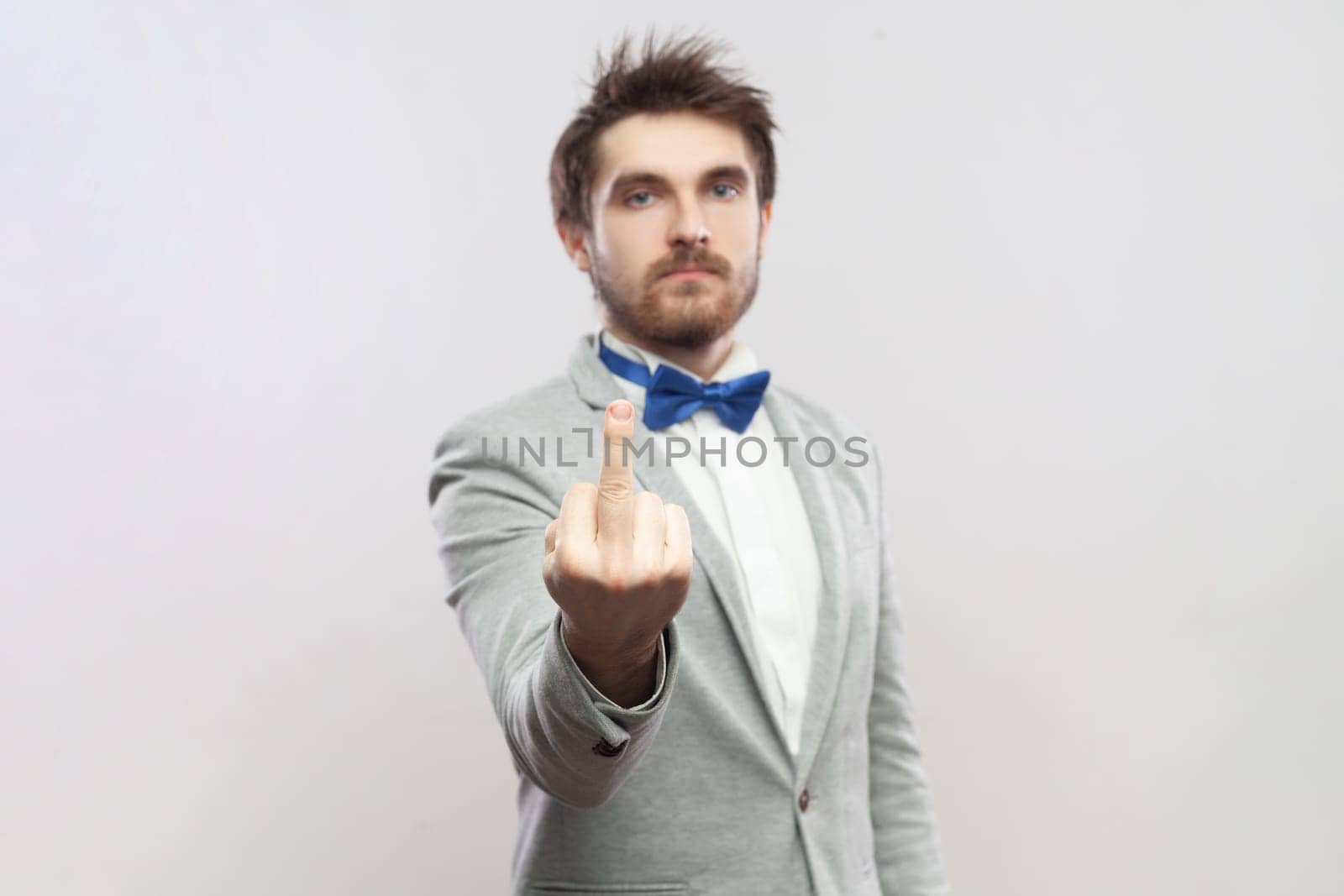 Portrait of rude impolite handsome bearded man standing showing middle finger, arguing with somebody, wearing grey suit and blue bow tie. Indoor studio shot isolated on gray background.