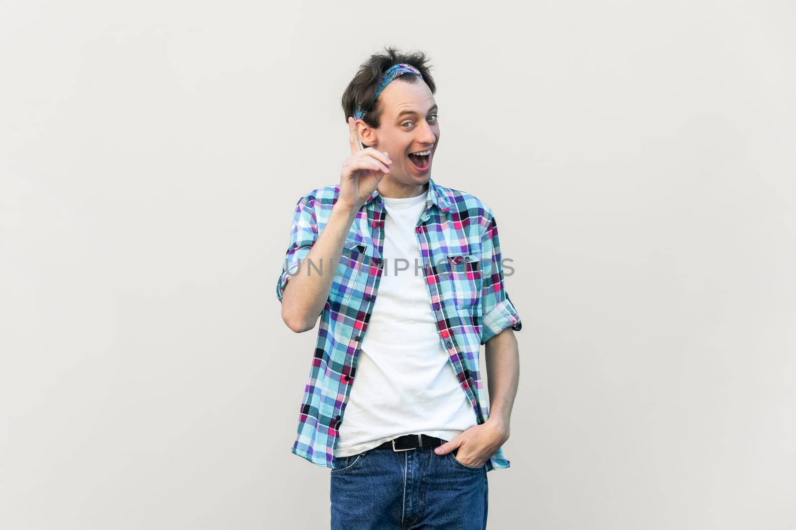 Optimistic glad excited man makes gesture with fore finger upwards, having great idea, smiling, wearing blue checkered shirt and headband. Indoor studio shot isolated on gray background.