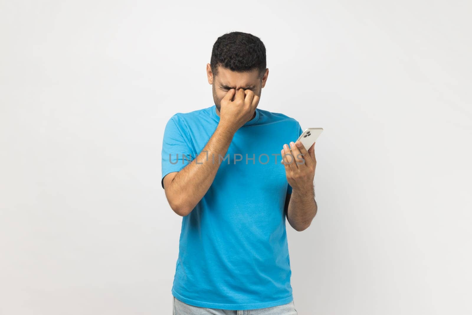Portrait of tired sad unshaven man wearing blue T- shirt standing holding smart phone in hands, feels pain in his eyes, looking at screen long hours. Indoor studio shot isolated on gray background.