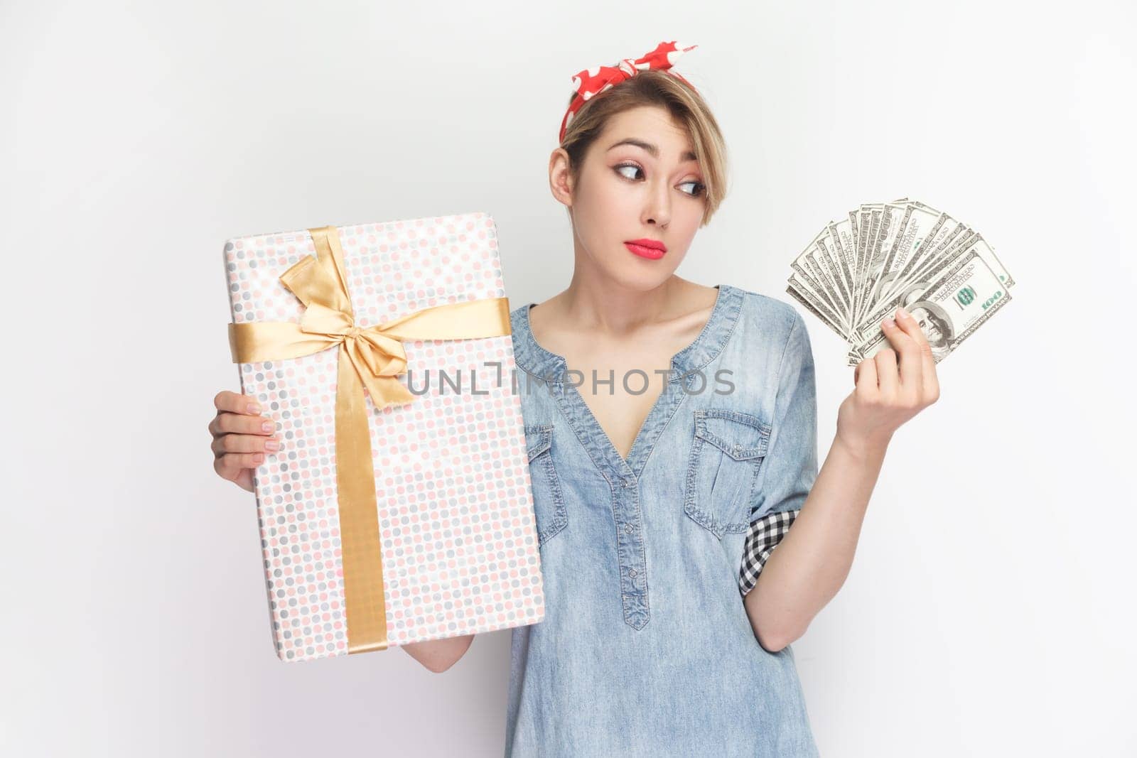 Portrait of confused puzzled uncertain blonde woman wearing blue denim shirt and red headband standing holding present box and dollar banknotes. Indoor studio shot isolated on gray background.