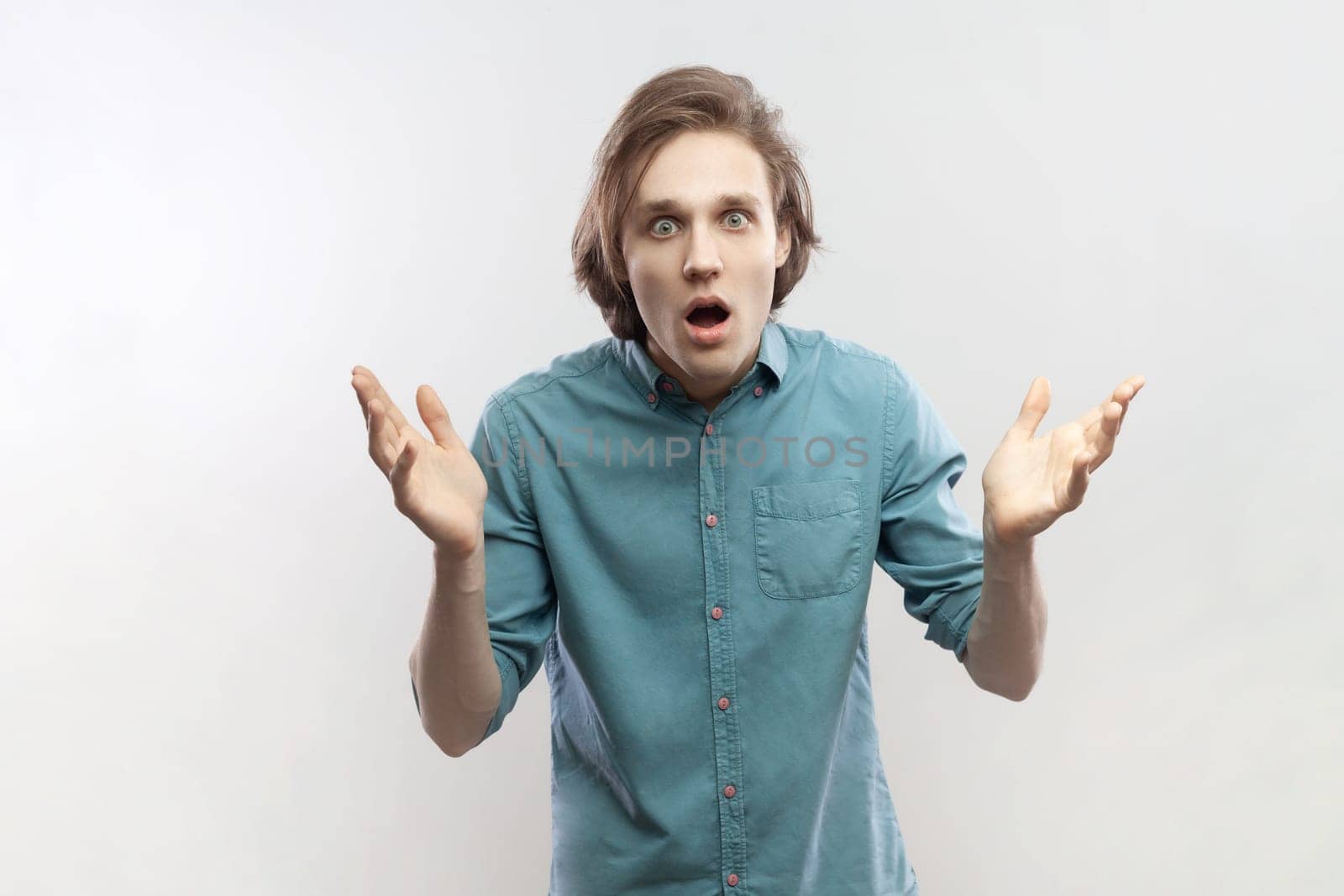 Portrait of shocked excited worried young man raised his arms, looking at camera, sees something unbelievable, saying wow, wearing blue shirt. Indoor studio shot isolated on gray background.