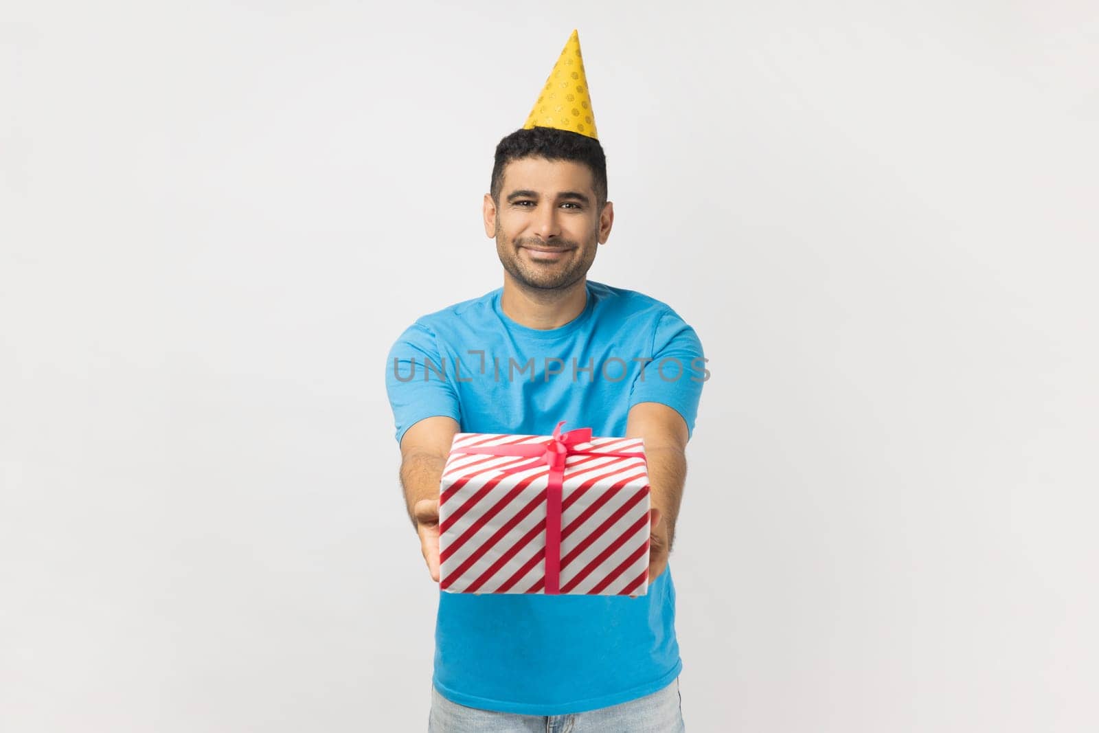 Portrait of cute attractive positive unshaven man wearing blue T- shirt and yellow party cone giving present box, having fun at birthday party. Indoor studio shot isolated on gray background.