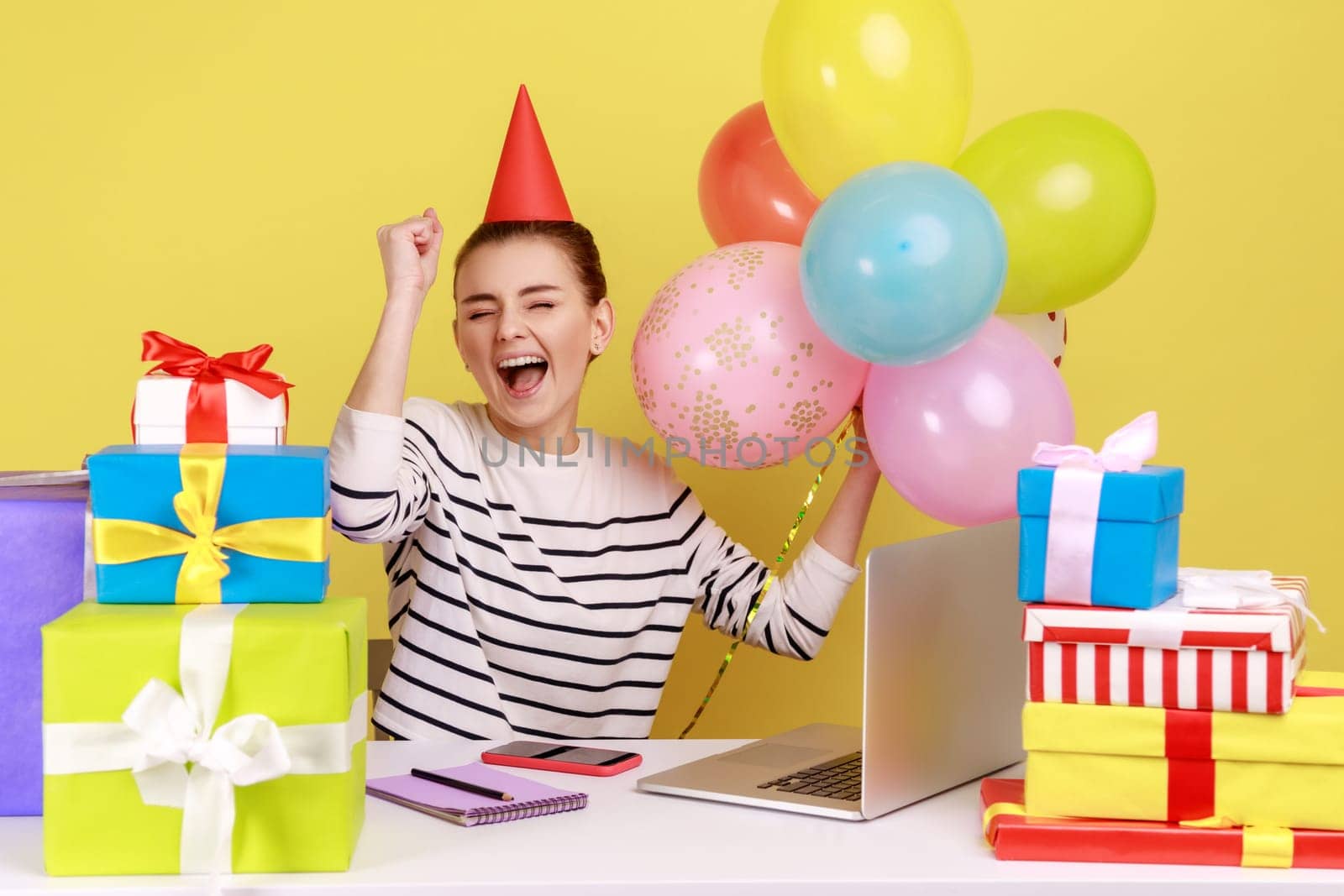 Excited woman office employee with party cone sitting workplace surrounded by gift boxes and balloons, shouting for joy, celebrating birthday. Indoor studio studio shot isolated on yellow background.