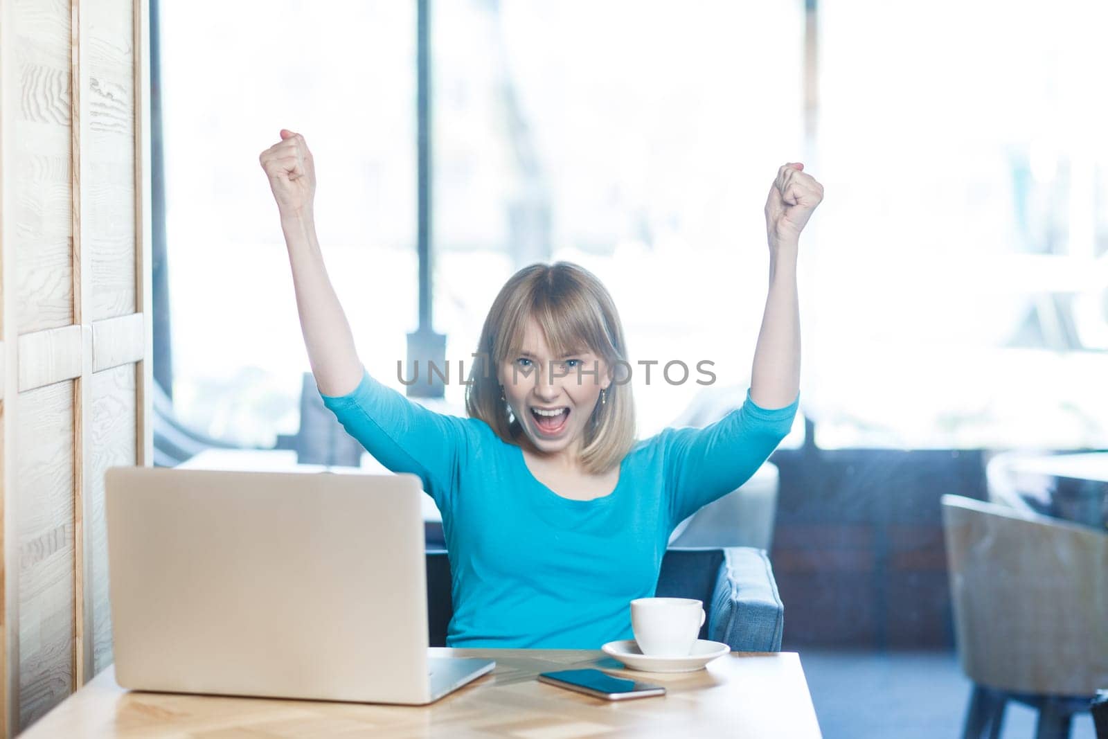 Extremely happy overjoyed young woman with blonde hair in blue shirt working on laptop, raised her clenched fists, celebrating finishing project. Indoor shot in cafe with big window on background.