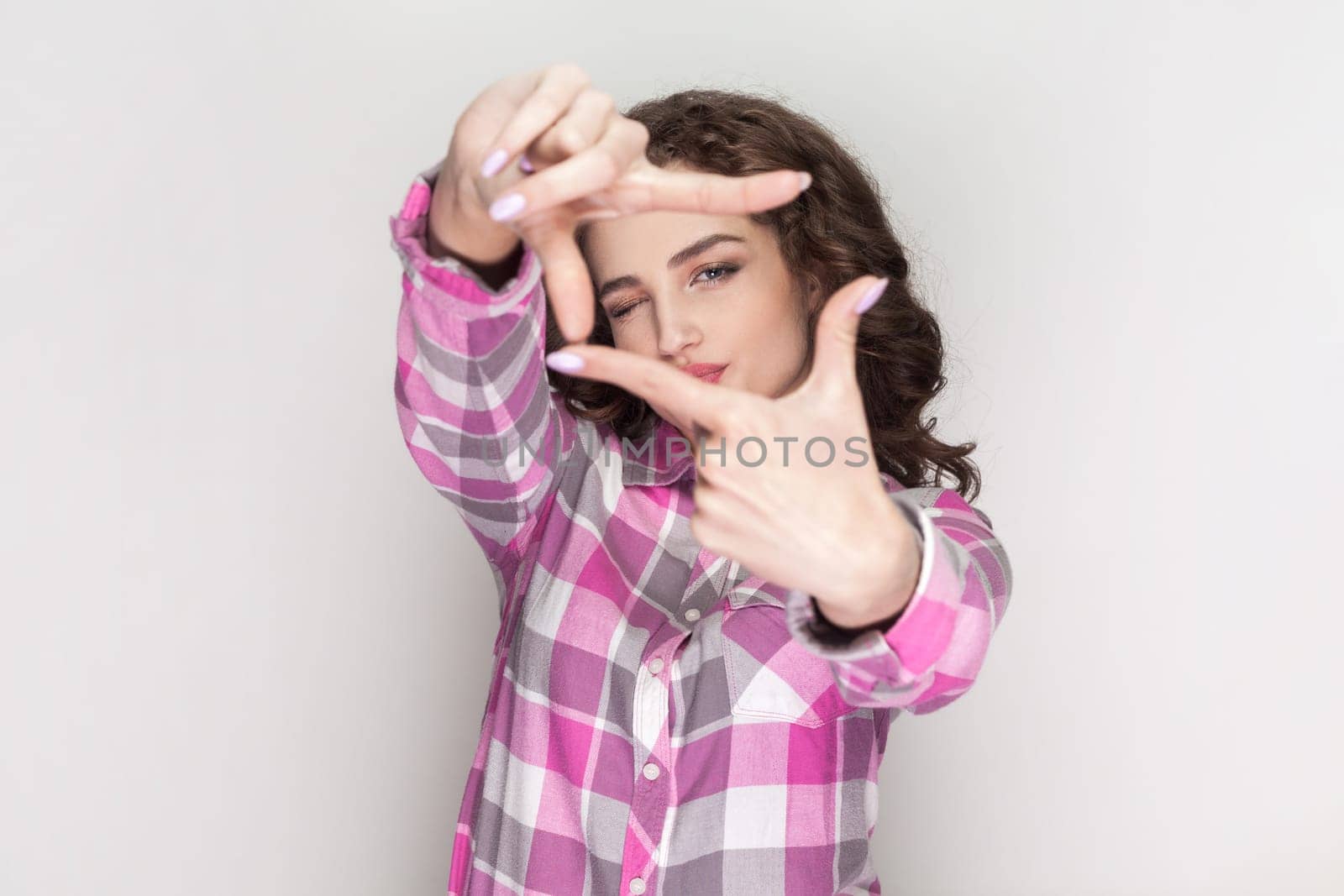 Happy ambitious woman with curly hair aims only success, makes hand frames searches perfect angle smiles broadly, gazes at camera through hands. Indoor studio shot isolated on gray background.