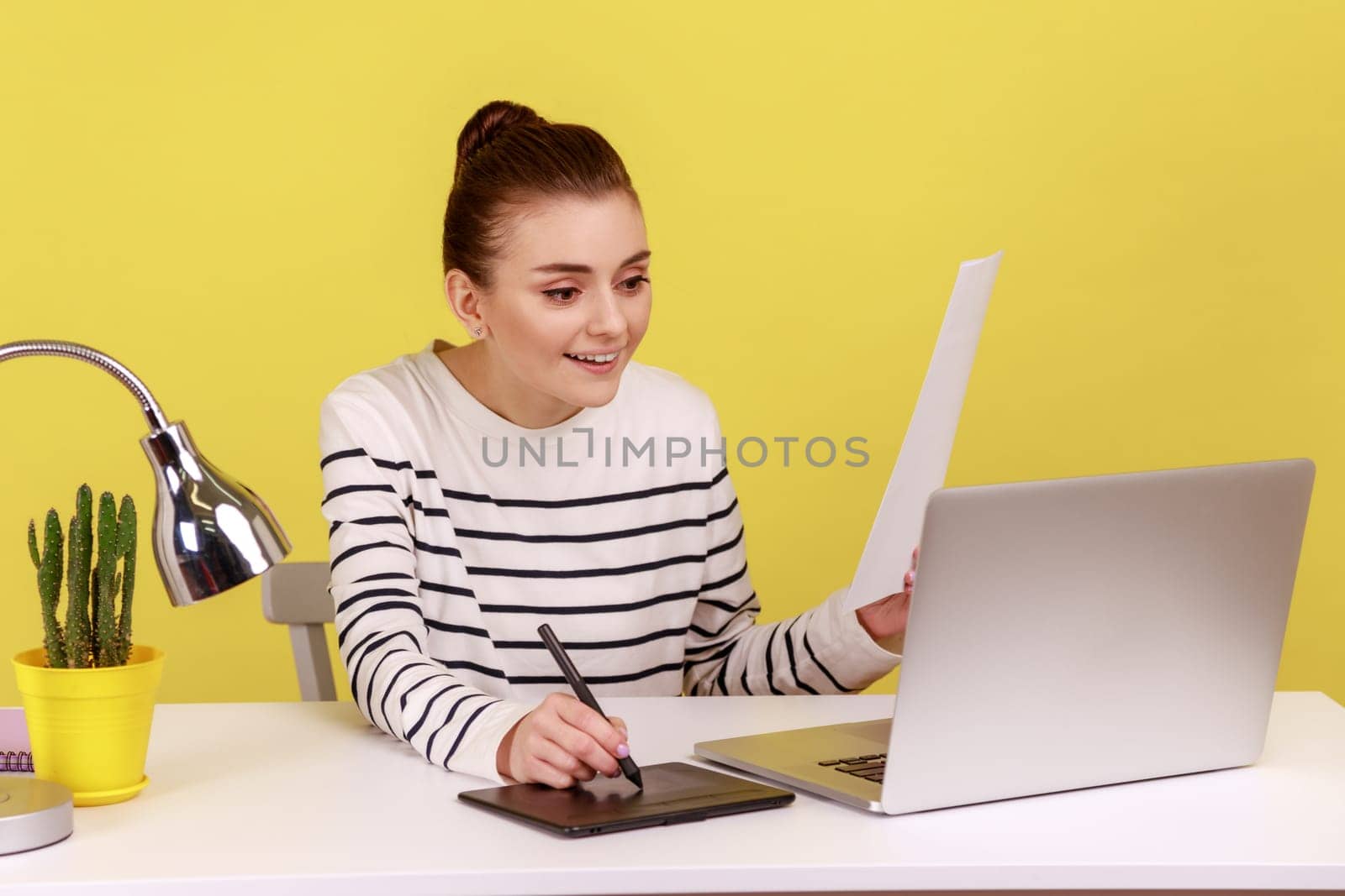 Portrait of smiling hard working woman designer drawing on professional tablet looking at laptop display, holding paper document. Indoor studio studio shot isolated on yellow background.