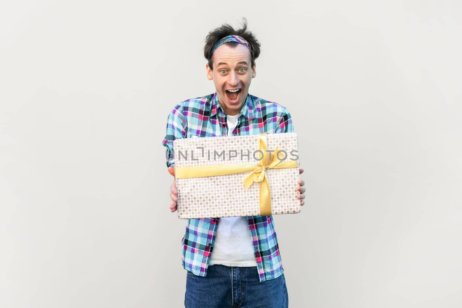 Excited man giving present box to his friend, being in good festive mood, screaming in astonishment, wearing blue checkered shirt and headband. Indoor studio shot isolated on gray background.