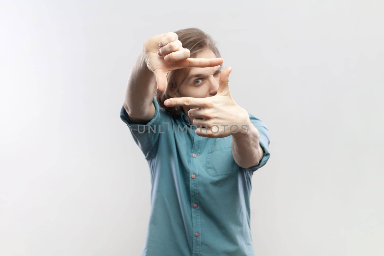 Portrait of man looking through frame of fingers gesturing photo, zooming focusing to camera, observing with serious expression, wearing blue shirt. Indoor studio shot isolated on gray background.