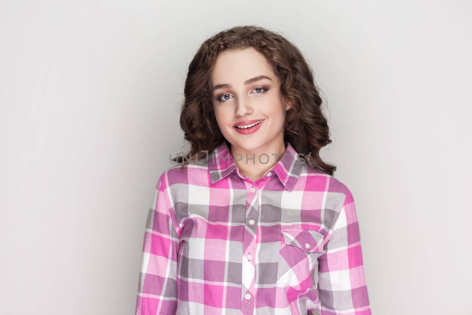 Cheerful woman with curly hair enjoys good day, looks unbothered and happy, gives sincere friendly smile to you, wearing pink checkered shirt. Indoor studio shot isolated on gray background.
