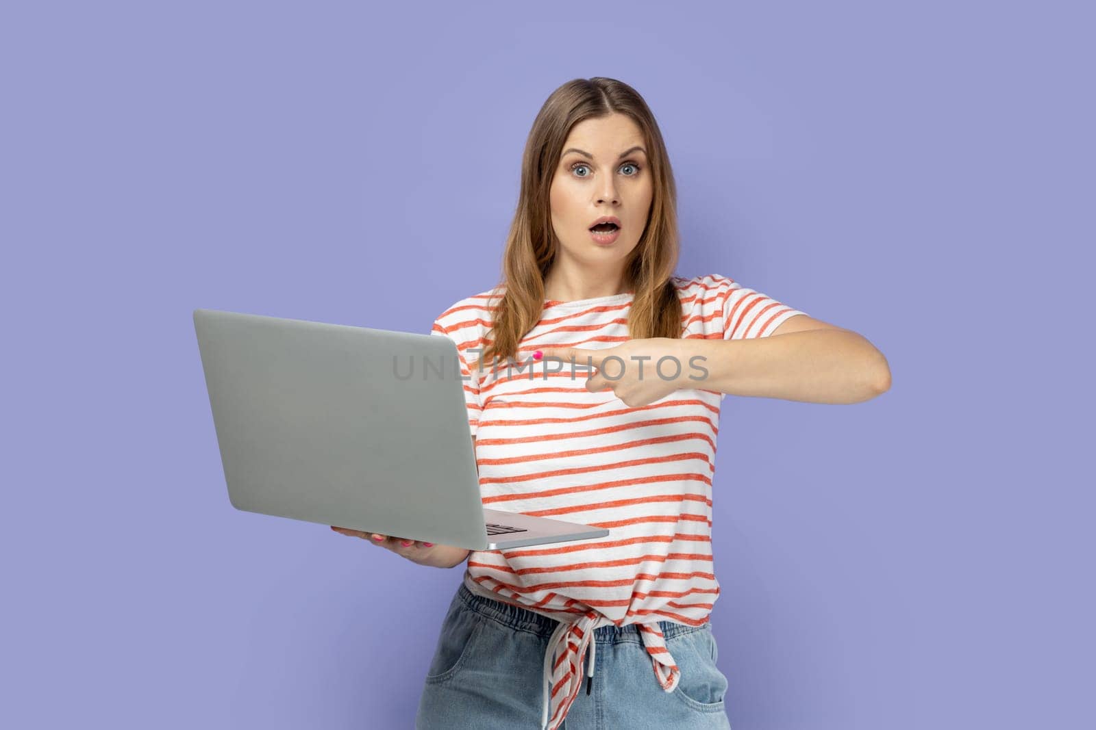 Portrait of shocked astonished blond woman wearing striped T-shirt working on laptop, looking at camera and pointing at display with finger. Indoor studio shot isolated on purple background.