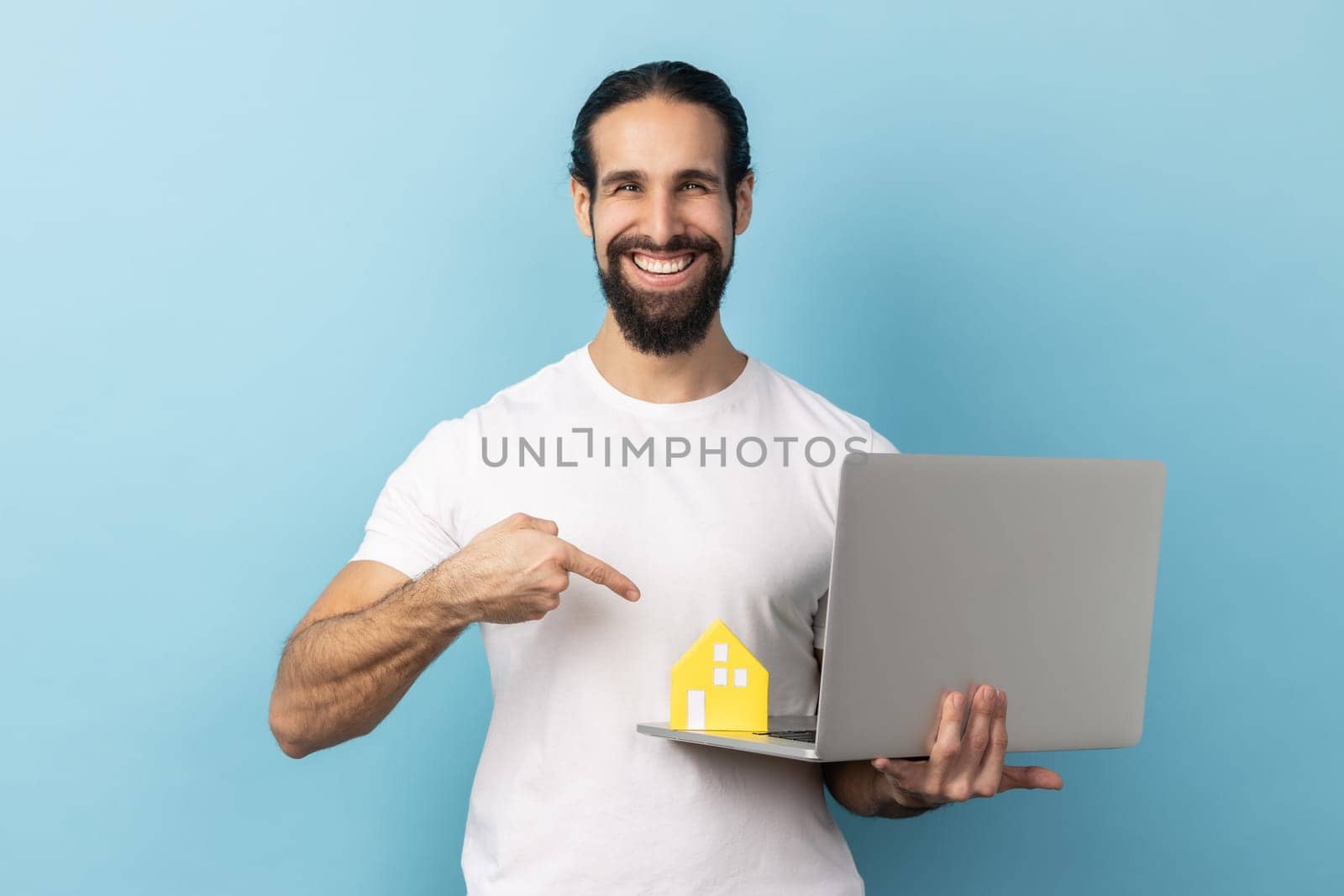 Portrait of happy man with beard wearing white T-shirt standing with paper and laptop, pointing at screen, helping with rent, working on project online. Indoor studio shot isolated on blue background.
