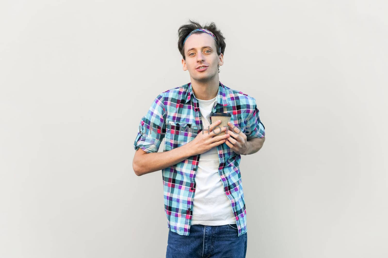 Smiling handsome man standing holding takeaway coffee in front of chest, looking at camera, love coffee, wearing blue checkered shirt and headband. Indoor studio shot isolated on gray background.