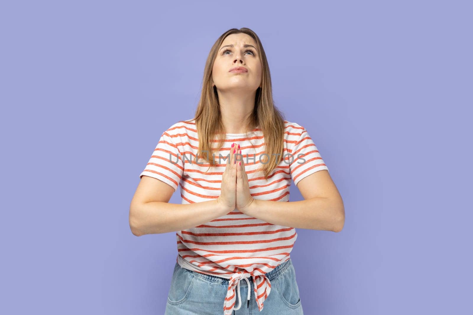 Portrait of attractive blond woman wearing striped T-shirt raising hands in prayer, looking up with hopeful pleading eyes, praying. Indoor studio shot isolated on purple background.