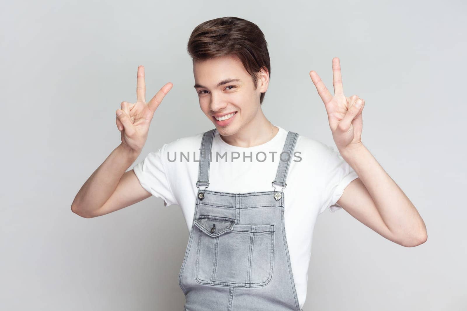 Portrait of young brunette man standing makes peace gesture, shows v sign, looking at camera with toothy smile, wearing denim overalls. Indoor studio shot isolated on gray background.