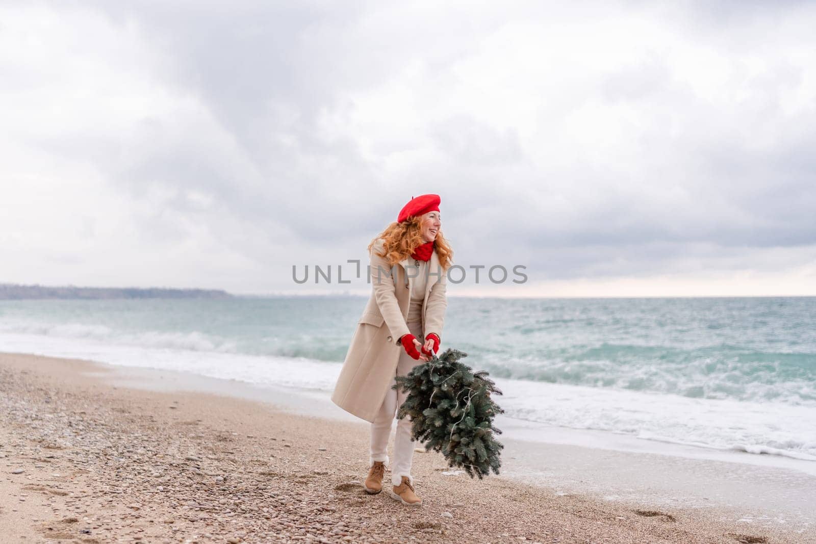 Redhead woman Christmas tree sea. Christmas portrait of a happy redhead woman walking along the beach and holding a Christmas tree in her hands. She is dressed in a light coat and a red beret