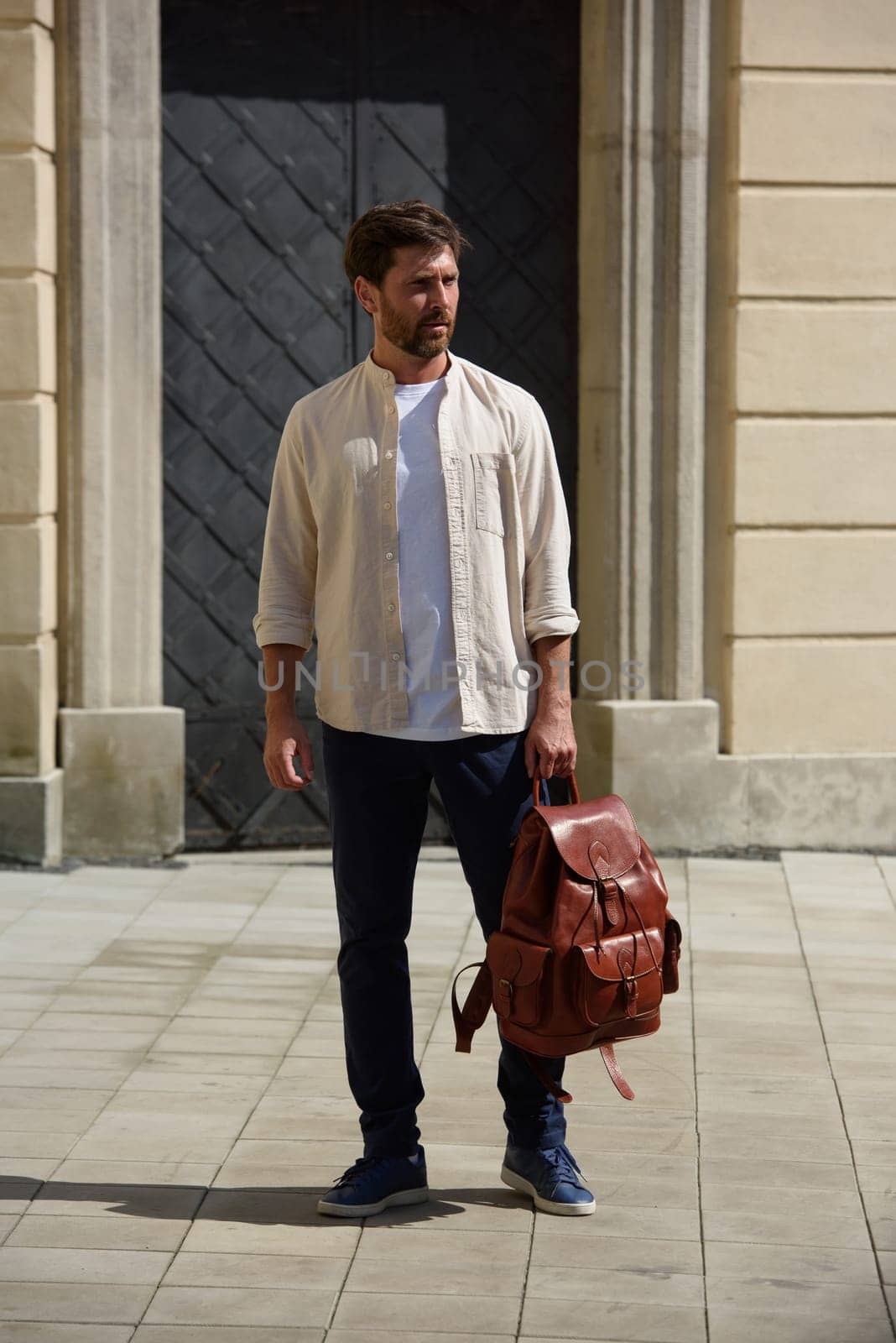 Handsome businessman walking on the street, with luxury leather backpack. Man wearing blue pants, sneakers, white t-shirt and beige shirt