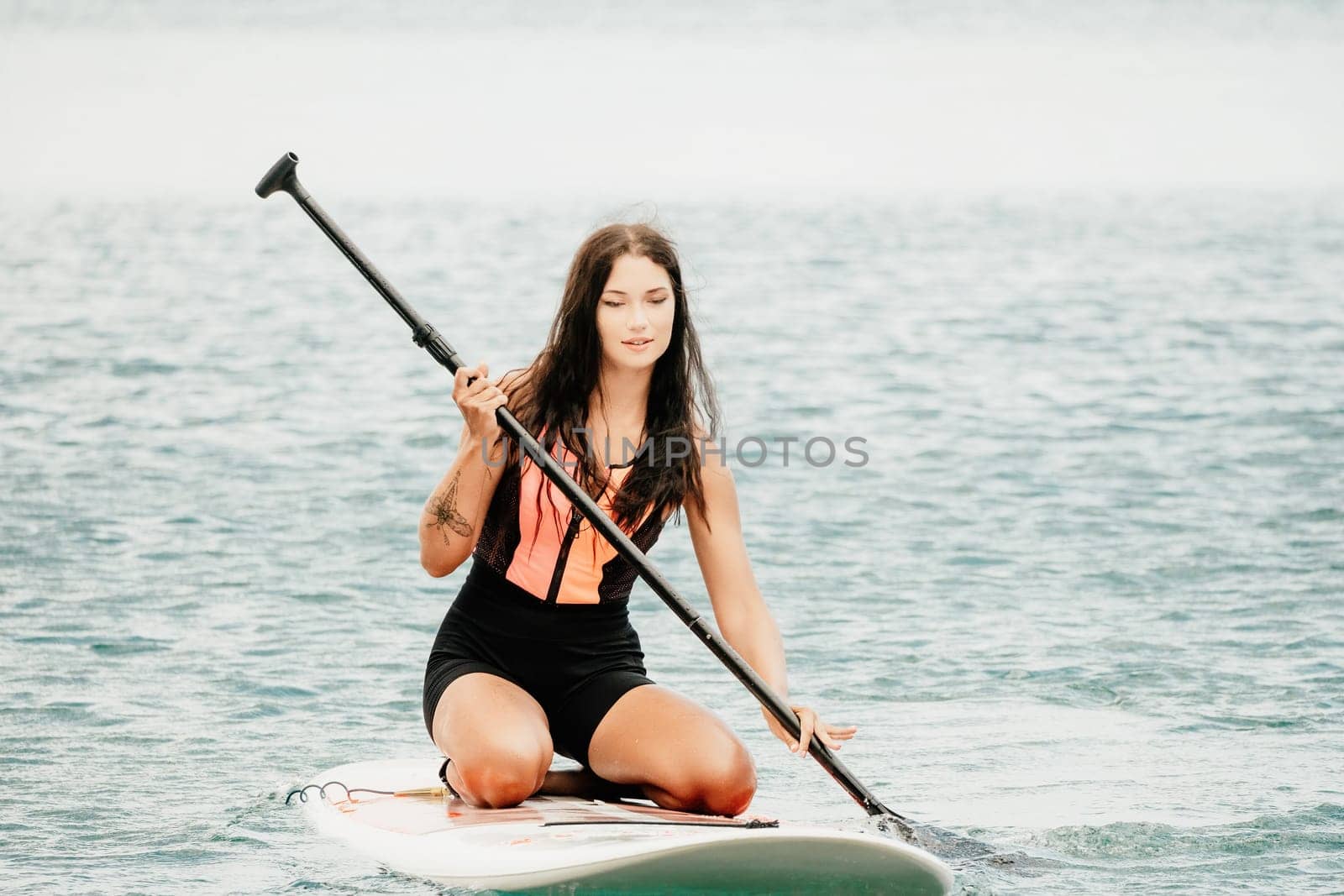 Close up shot of beautiful young caucasian woman with black hair and freckles looking at camera and smiling. Cute woman portrait in a pink bikini posing on sup board in the sea by panophotograph