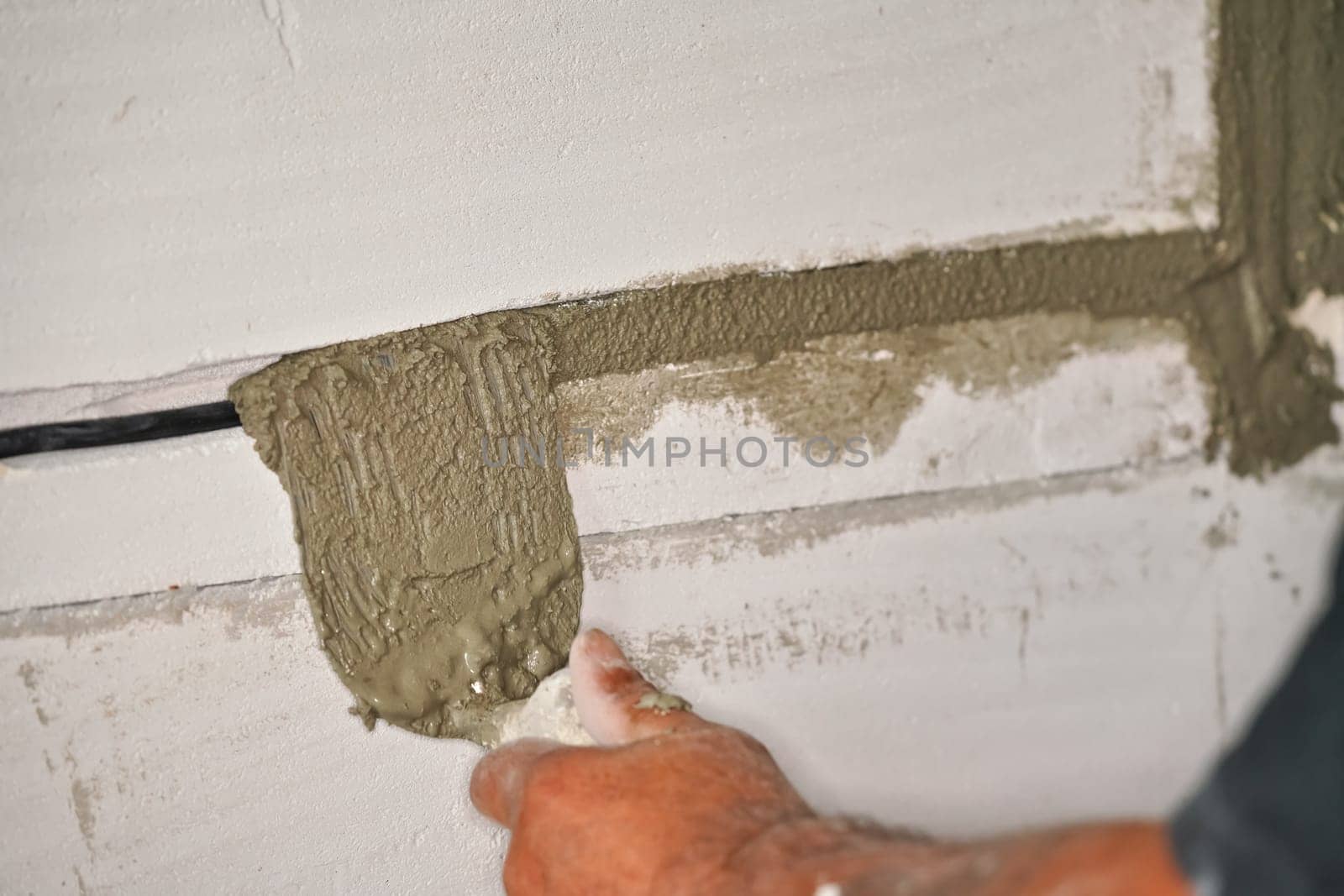 Construction worker securing electric sockets groove installed on bare wall with mortar cement paste - filling holes using small scoop, detail to hands only by Ivanko