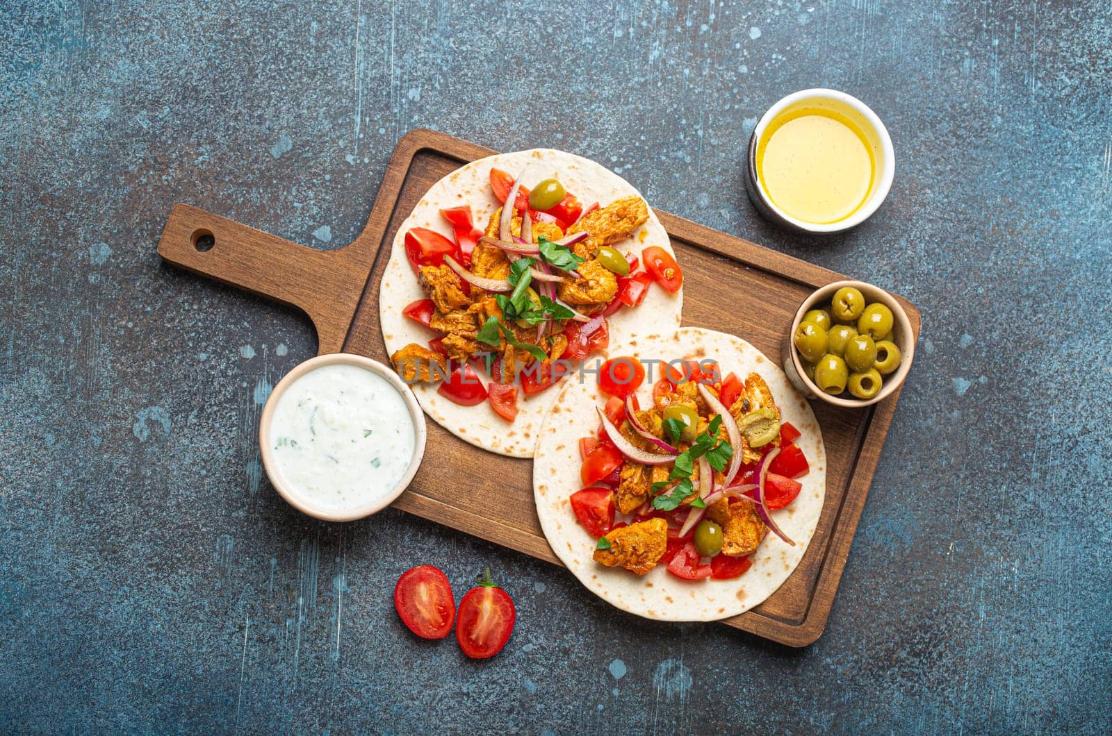 Cooking Traditional Greek Dish Gyros: Pita bread with vegetables, meat, herbs, olives on rustic wooden cutting board with Tzatziki sauce, olive oil top view on dark blue stone background.