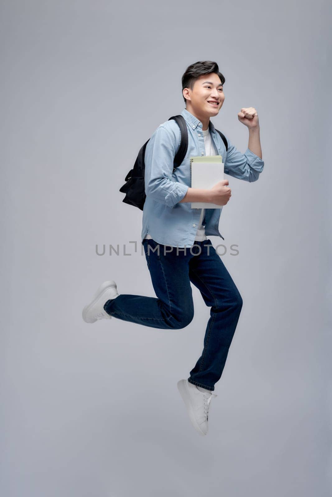 Full length portrait of a smiling young asian man carrying backpack isolated, holding books, jumping by makidotvn