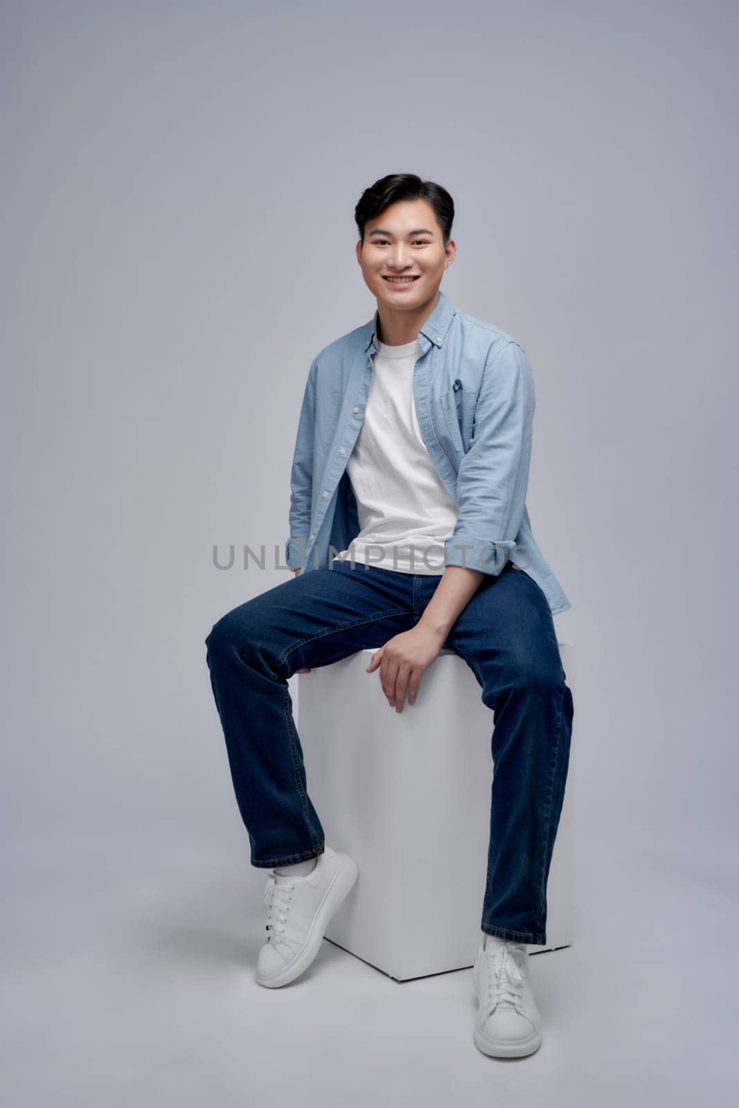 Young asian man posing while seated on box looking away from the camera in studio background