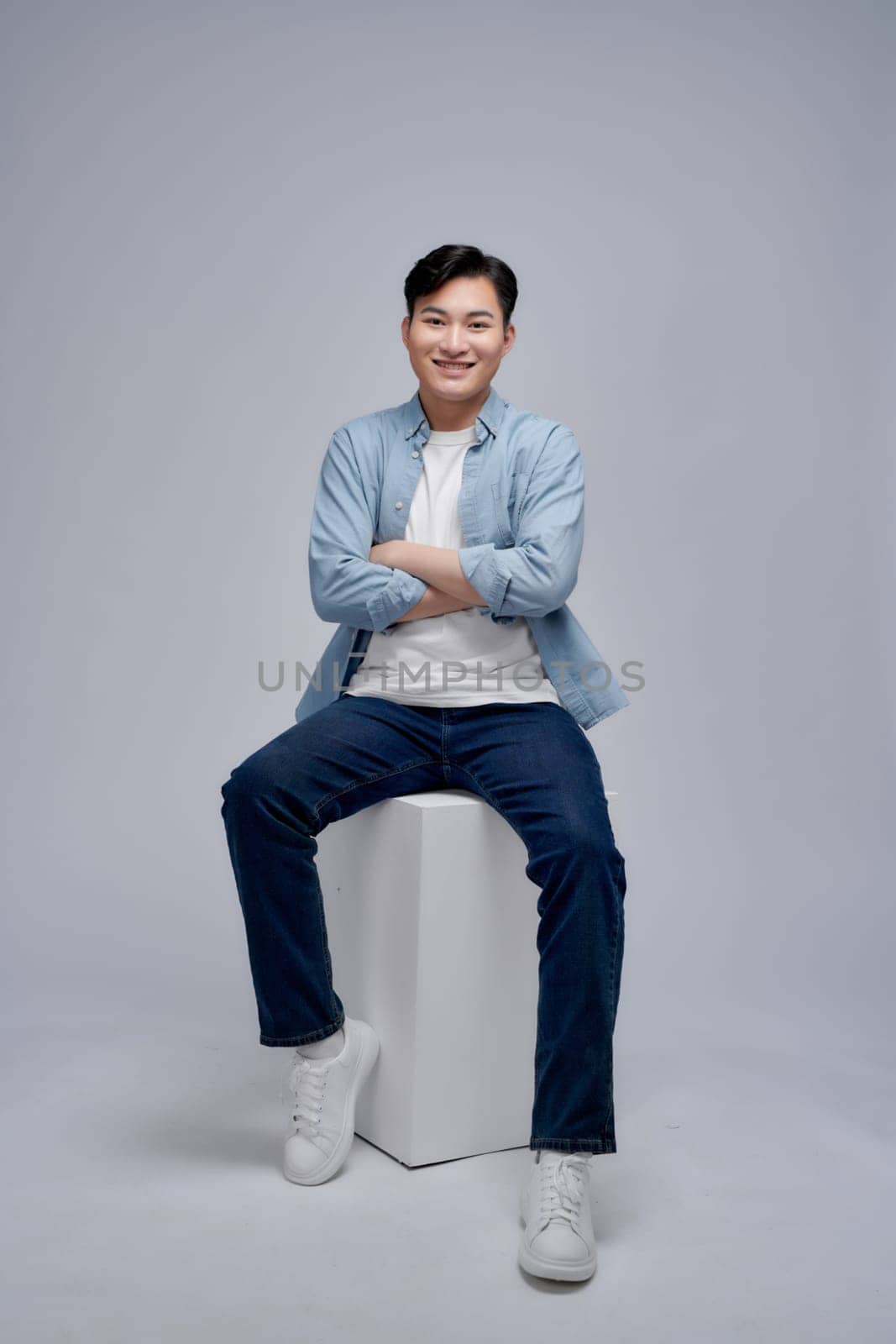 Full length of handsome young man looking at camera with smile while sitting against white background by makidotvn