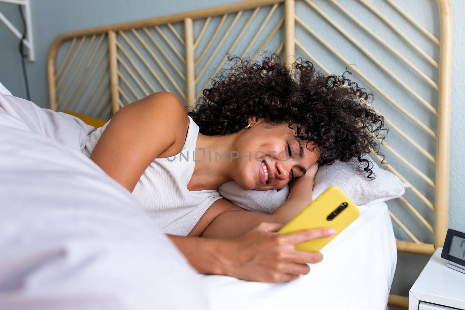 Happy woman using mobile phone in bed. Smiling black latina female looking mobile phone in the morning after waking up. by Hoverstock