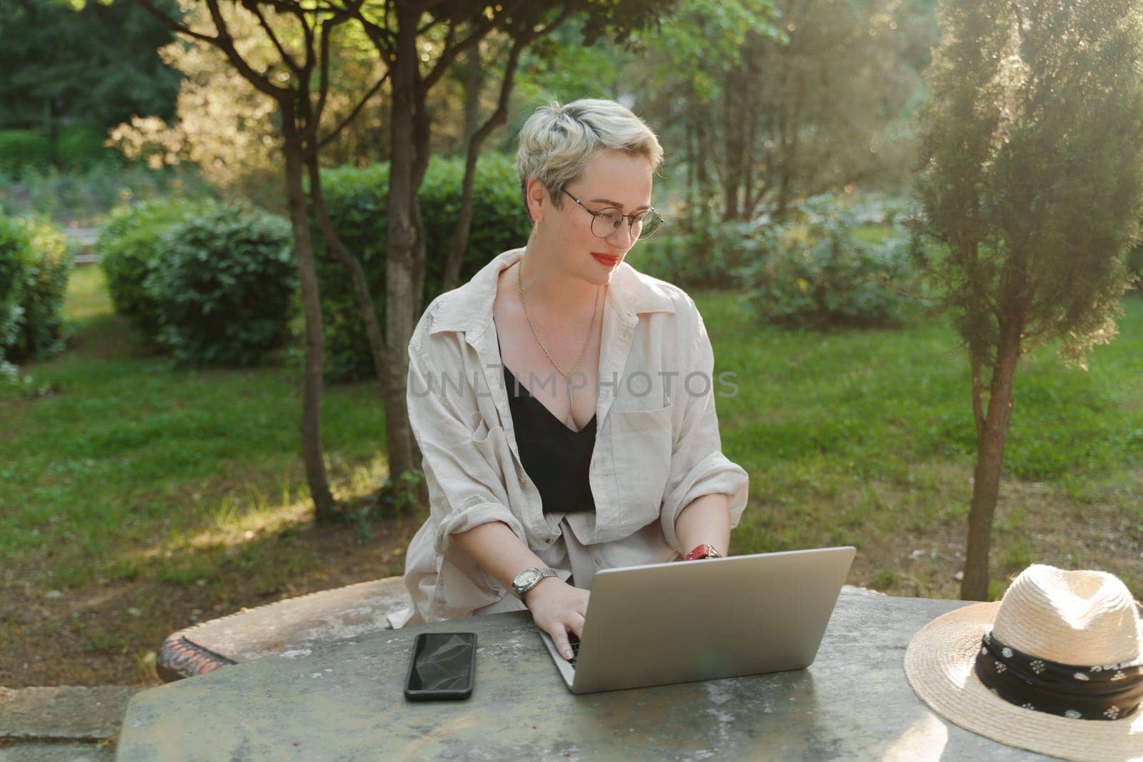 woman freelancer in glasses works at a computer at a white table in nature and spends her day productively.