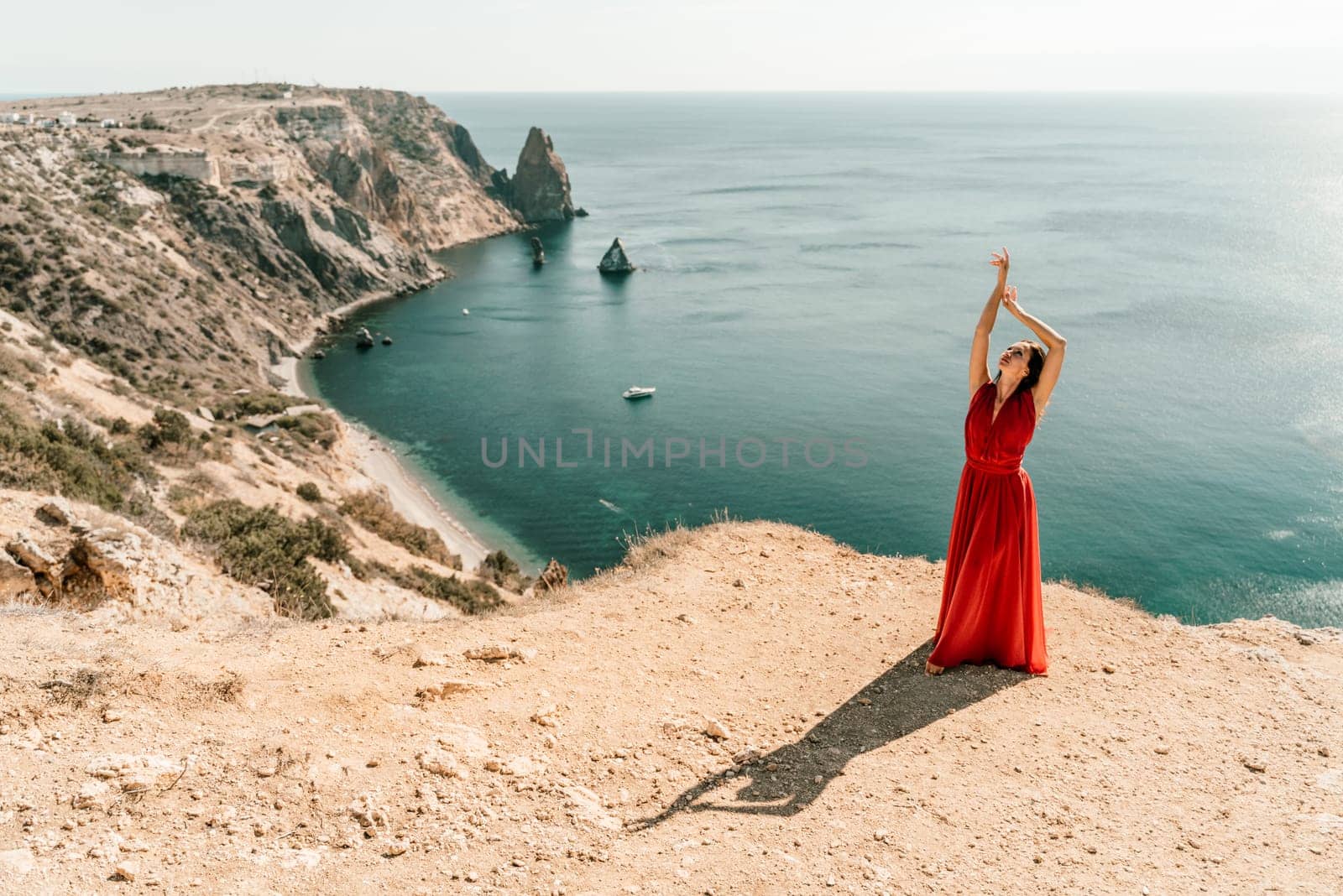 Woman red dress sea. posing on a rocky outcrop high above the sea. Girl on the nature on blue sky background. Fashion photo. by Matiunina
