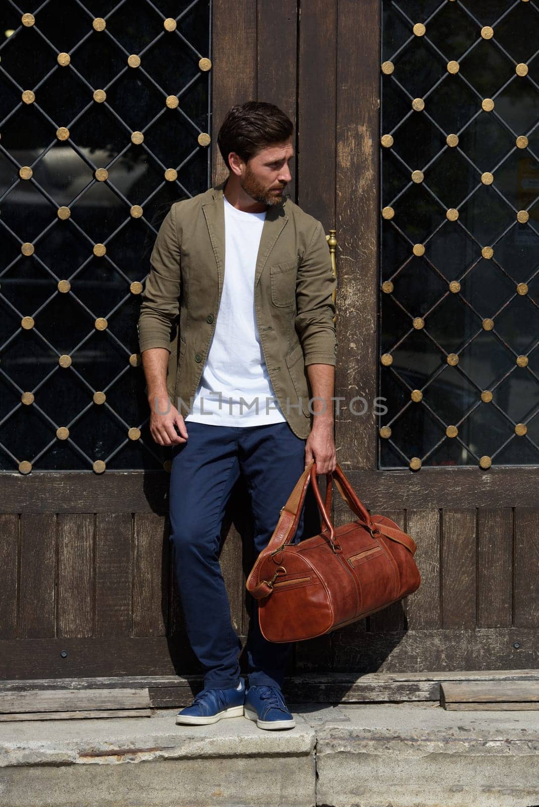 a man with a bag. black leather travel bag, street photo. man is wearing blue sneakers, jeans, a white T-shirt and a jacket.