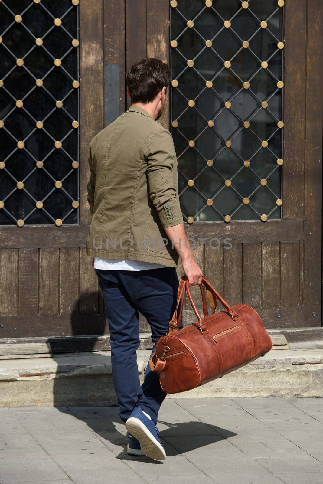 a man with a bag. black leather travel bag, street photo. man is wearing blue sneakers, jeans, a white T-shirt and a jacket.