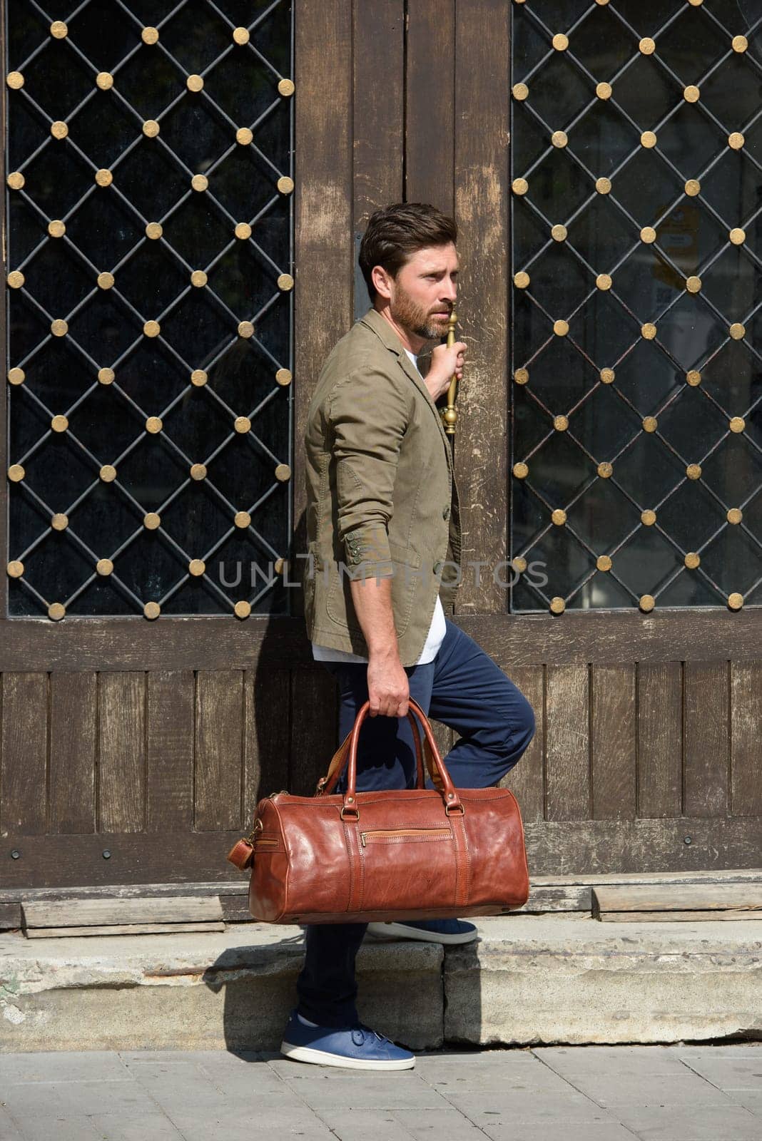 a man with a bag. black leather travel bag, street photo. man is wearing blue sneakers, jeans, a white T-shirt and a jacket by Ashtray25