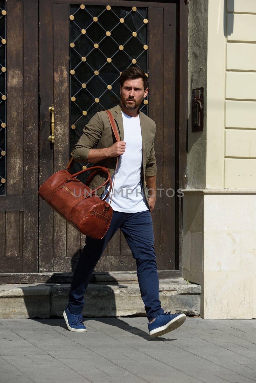 a man with a bag. black leather travel bag, street photo. man is wearing blue sneakers, jeans, a white T-shirt and a jacket.