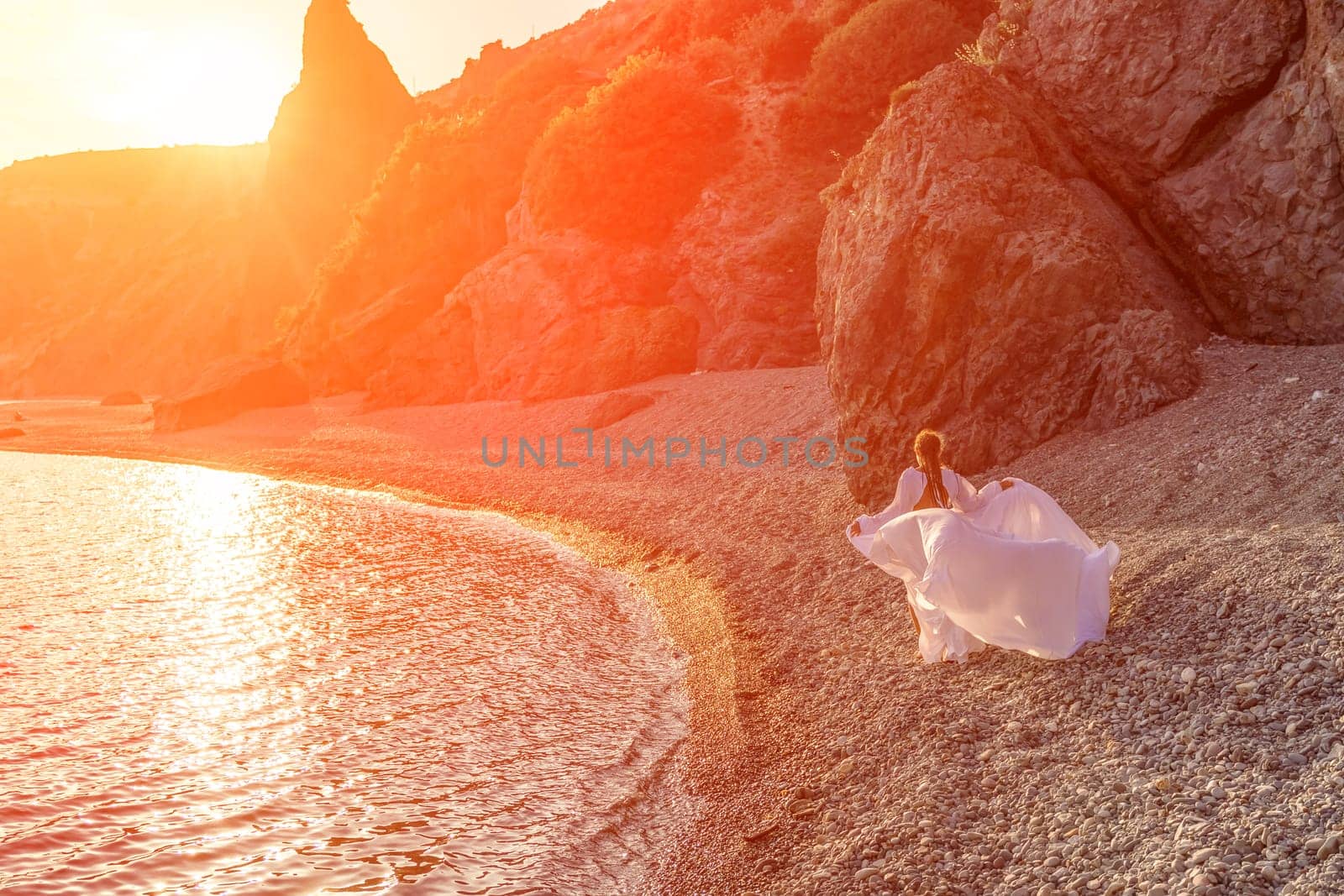 Mysterious woman silhouette long hair walks on the beach ocean water, sea nymph wind listens to the wave. Throws up a long white dress, a divine sunset. Artistic photo from the back without a face.