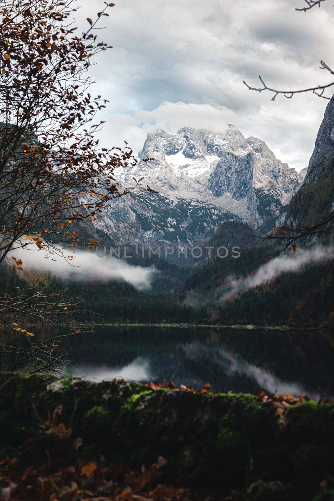 Autumn shot of a sunset at Lake Gosausee framed by orange and yellow trees against the backdrop of the Austrian Alps. The clouds lie low on the trees. There are yellowed leaves on the ground, the mountain and the sky are reflected in the lake.
