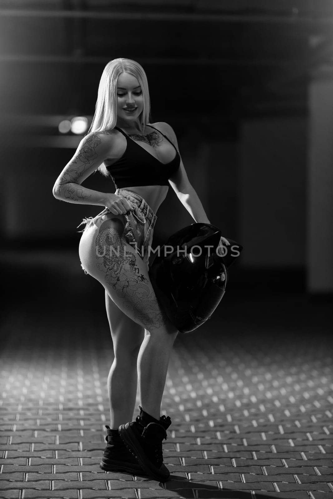 Sexy blonde woman model with silicone breasts and tattoos posing with motorcycle helmet in her hands, black and white photo