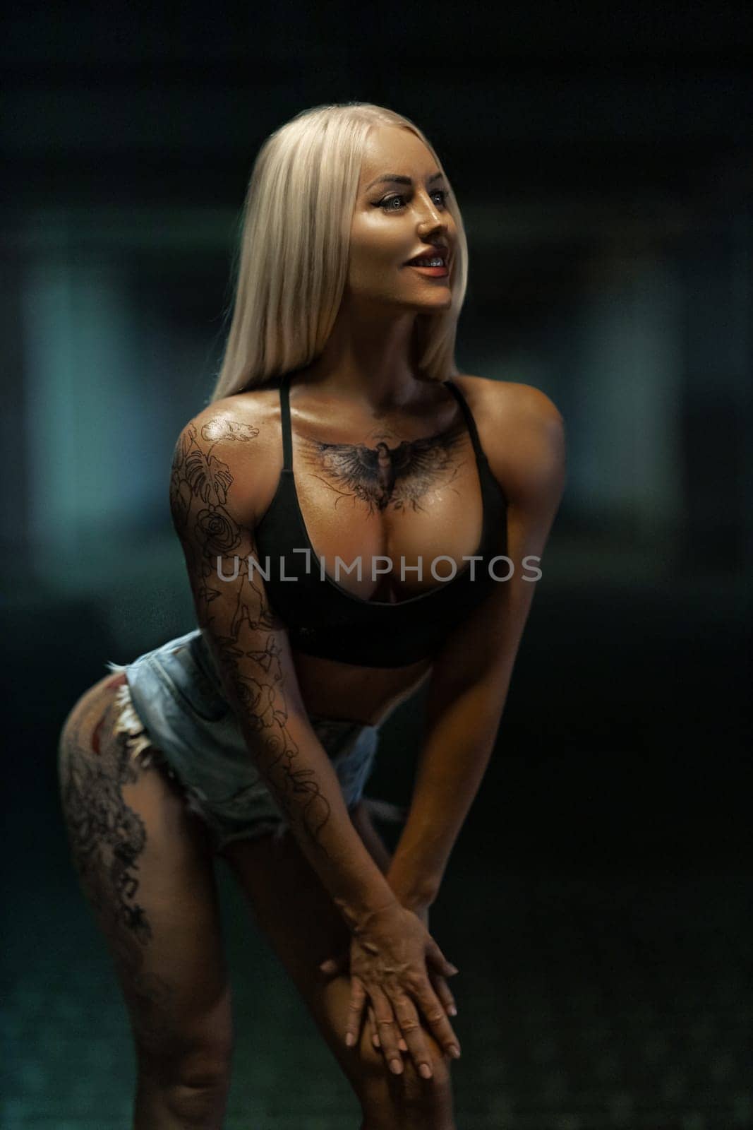 Sexy blonde woman fitness model in denim shorts and black top poses with both hands on her leg, showing off her beautiful silicone breasts with a bird tattoo