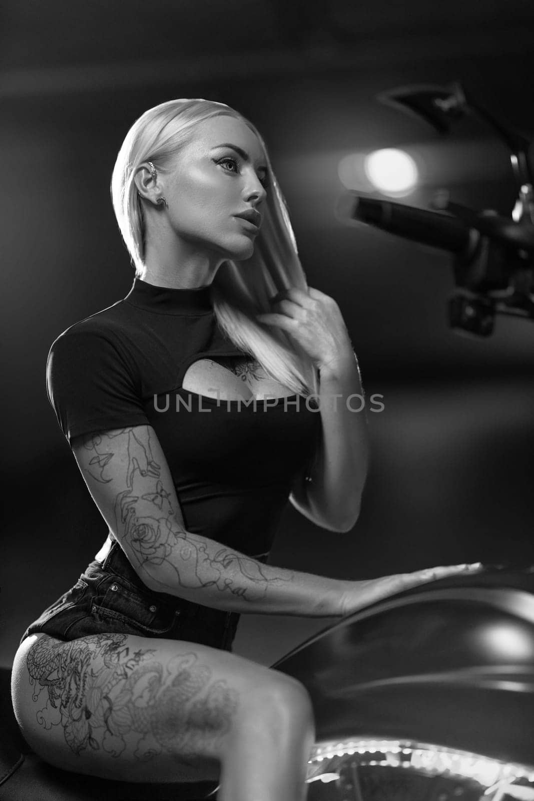 Sexy blonde woman with open neckline poses on motorcycle in the parking lot. Beautiful fitness model with tattoos sits on motorbike, black and white photo