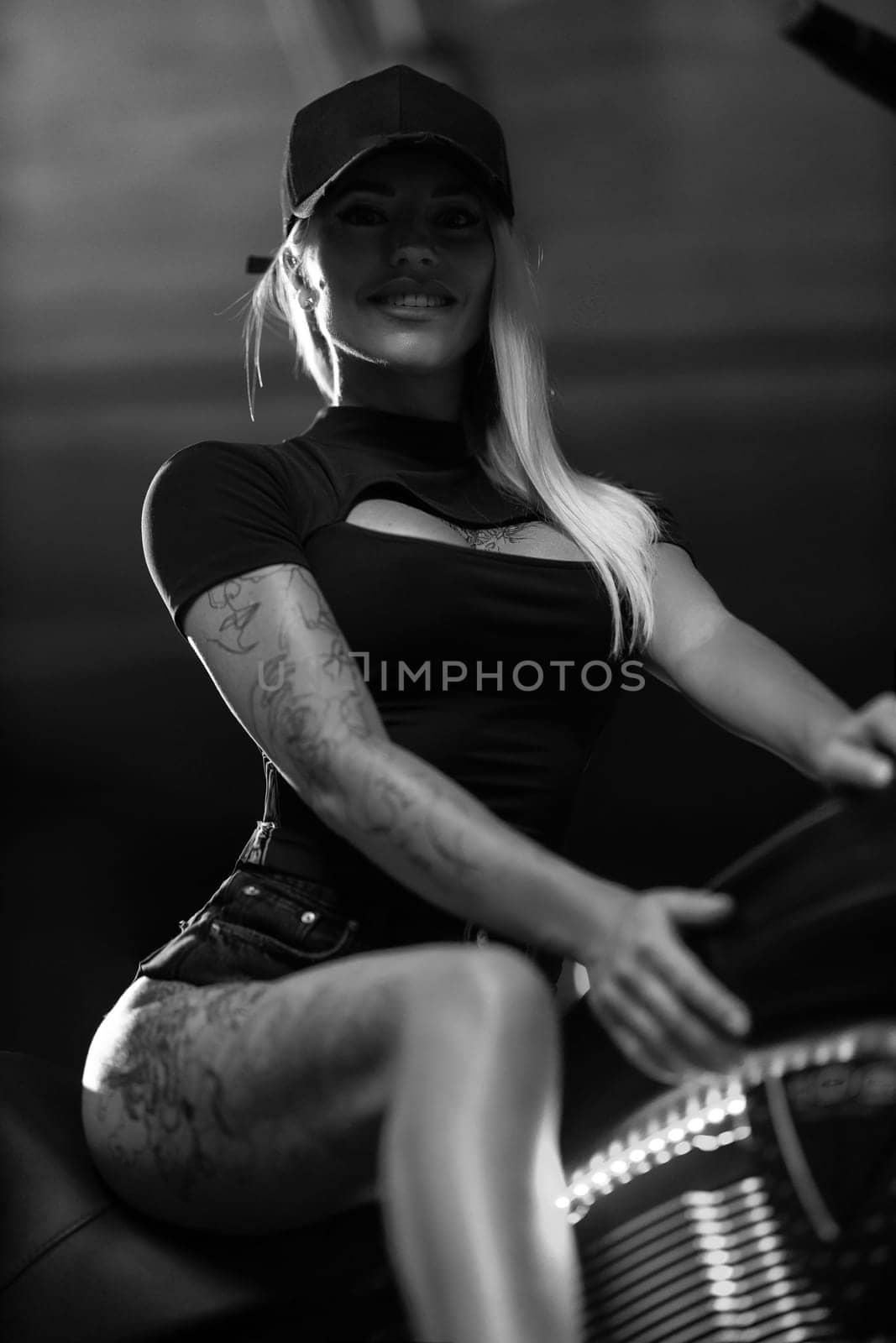 Sexy blonde woman with open neckline poses on motorcycle in the parking lot. Beautiful fitness model with tattoos sits on motorbike, black and white photo