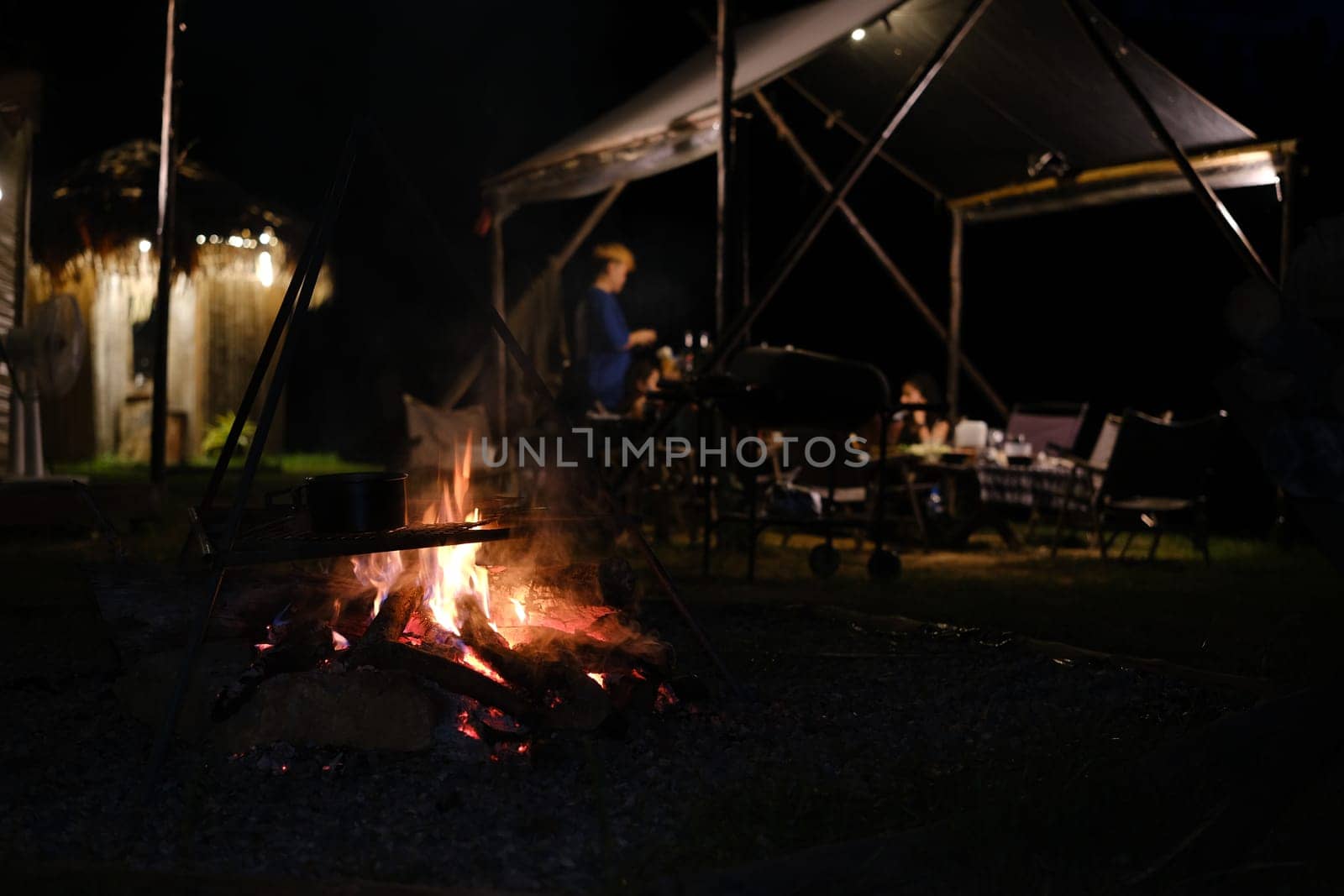 Bonfire near camping tent outdoors in evening and group of people sitting in background.