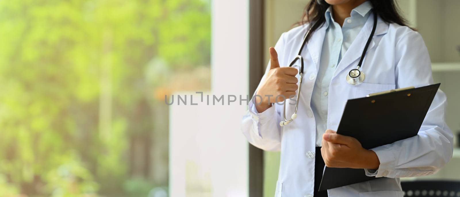 Doctor dressed in white overall with stethoscope holding clipboard and showing thumbs up.
