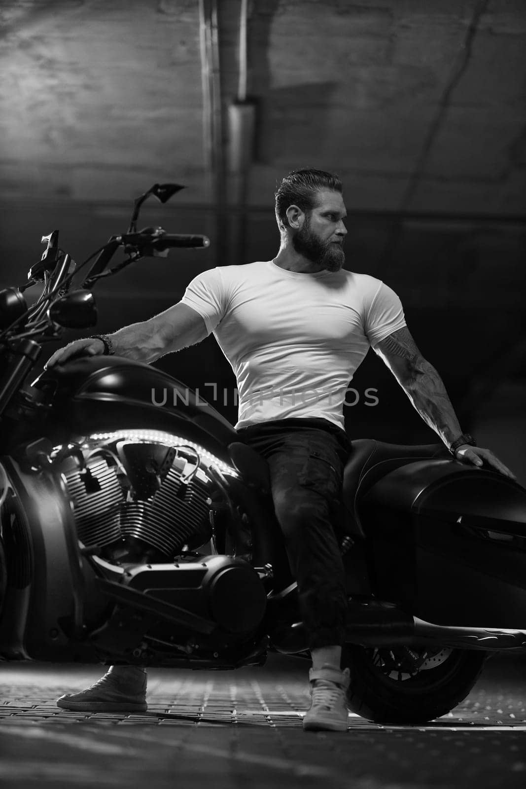 Sexy handsome attractive sporty muscular fitness model breaded biker in white tight tshirt sits on brutal beautiful motorcycle in the parking, black and white photo