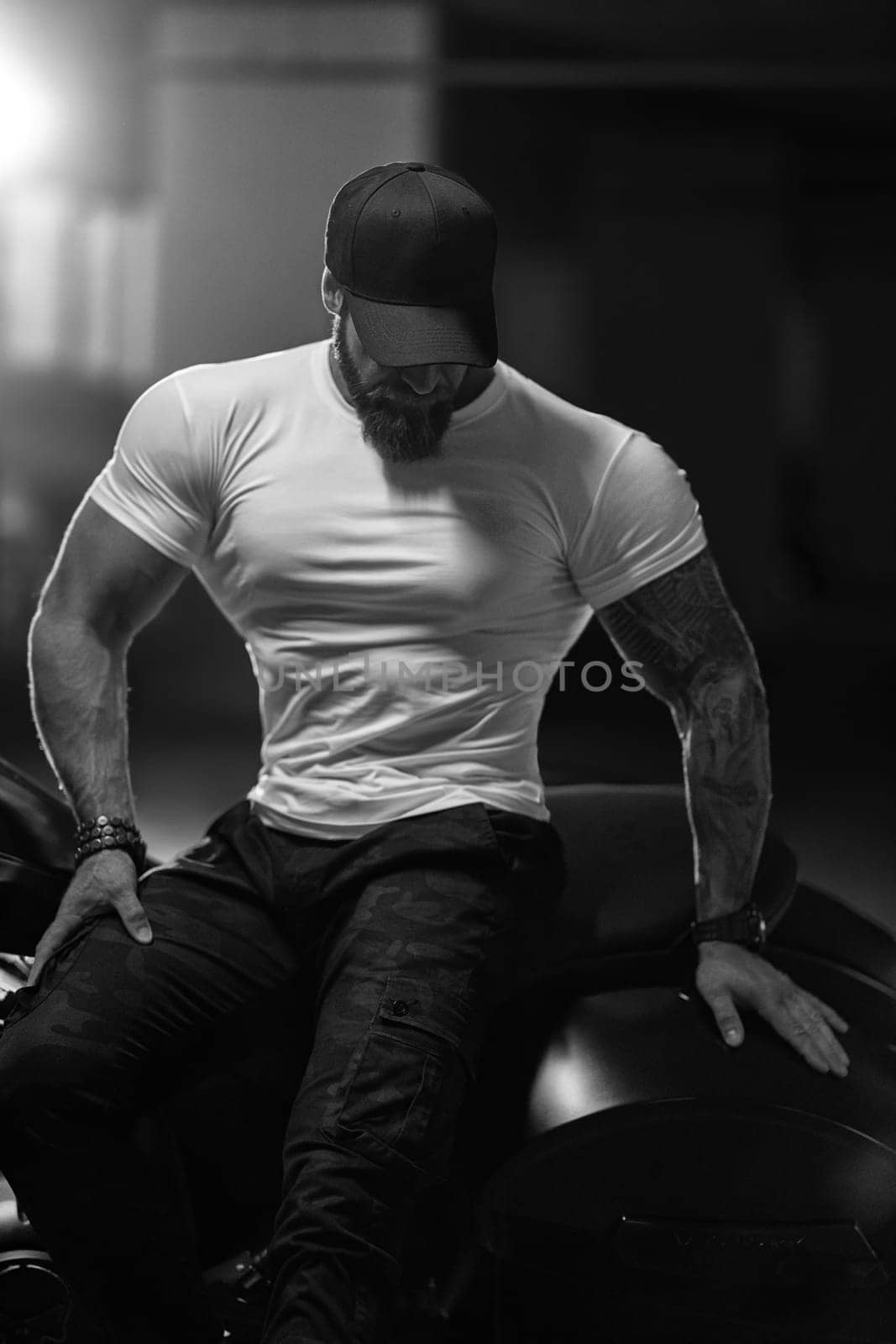 Sexy handsome attractive sporty muscular fitness model breaded biker in black cap and white tight tshirt sits on brutal beautiful motorcycle in the parking, black and white photo