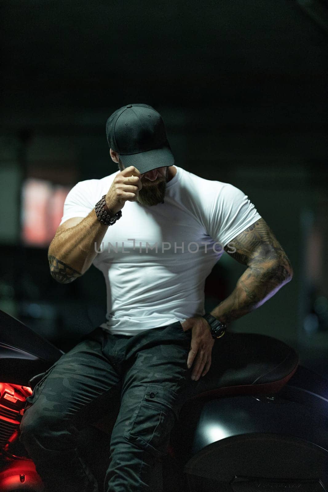 Sexy handsome attractive sporty muscular fitness model breaded biker in black cap and white tight tshirt sits on brutal beautiful motorcycle in the parking