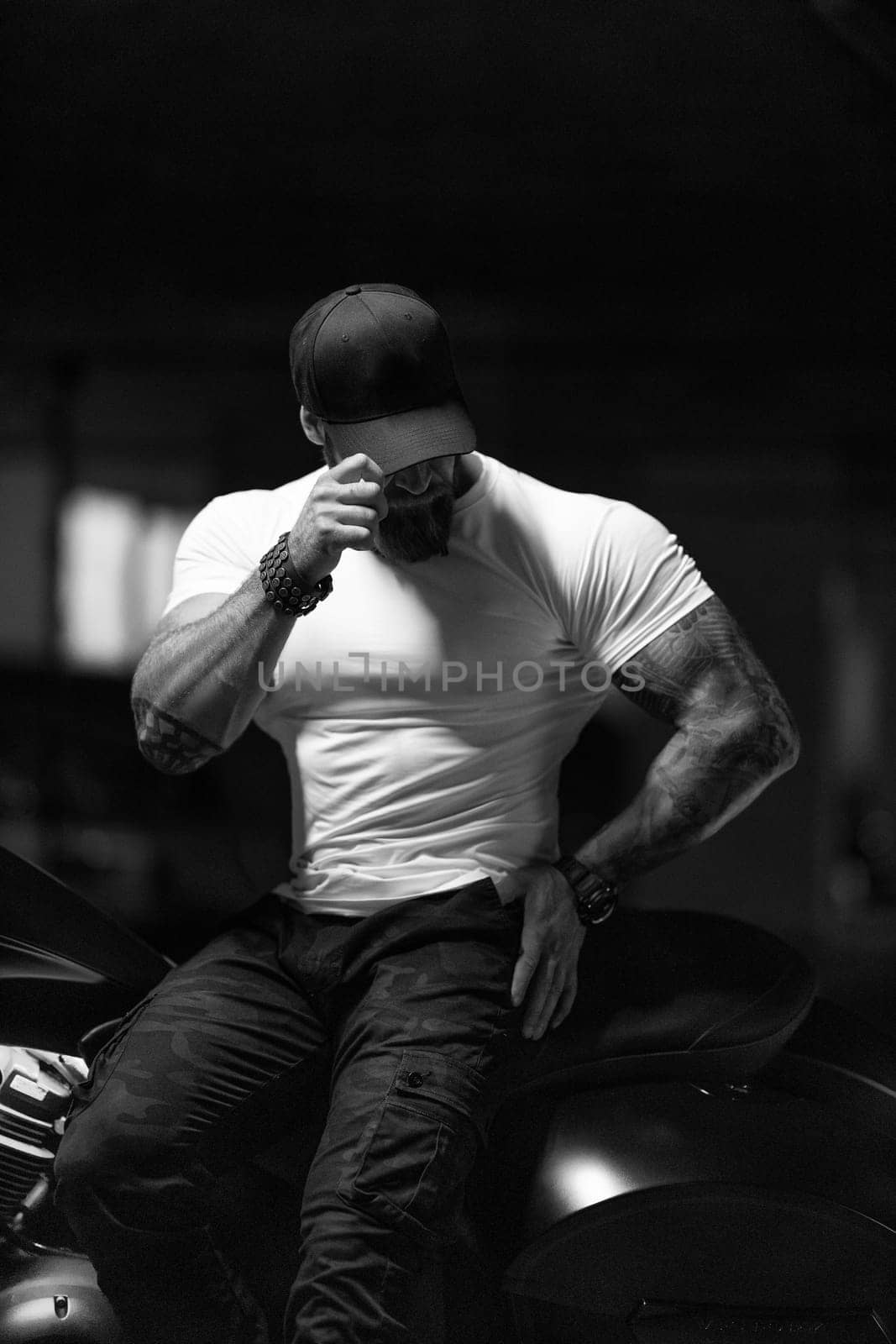 Sexy handsome attractive sporty muscular fitness model breaded biker in black cap and white tight tshirt sits on brutal beautiful motorcycle in the parking, black and white photo