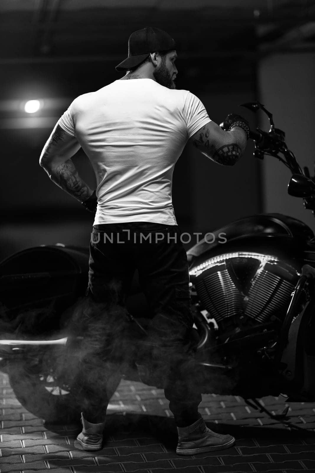 Sexy handsome attractive sporty muscular fitness model breaded biker with brutal beautiful motorcycle in the parking posing with a motorbike with his back to the frame, black and white photo