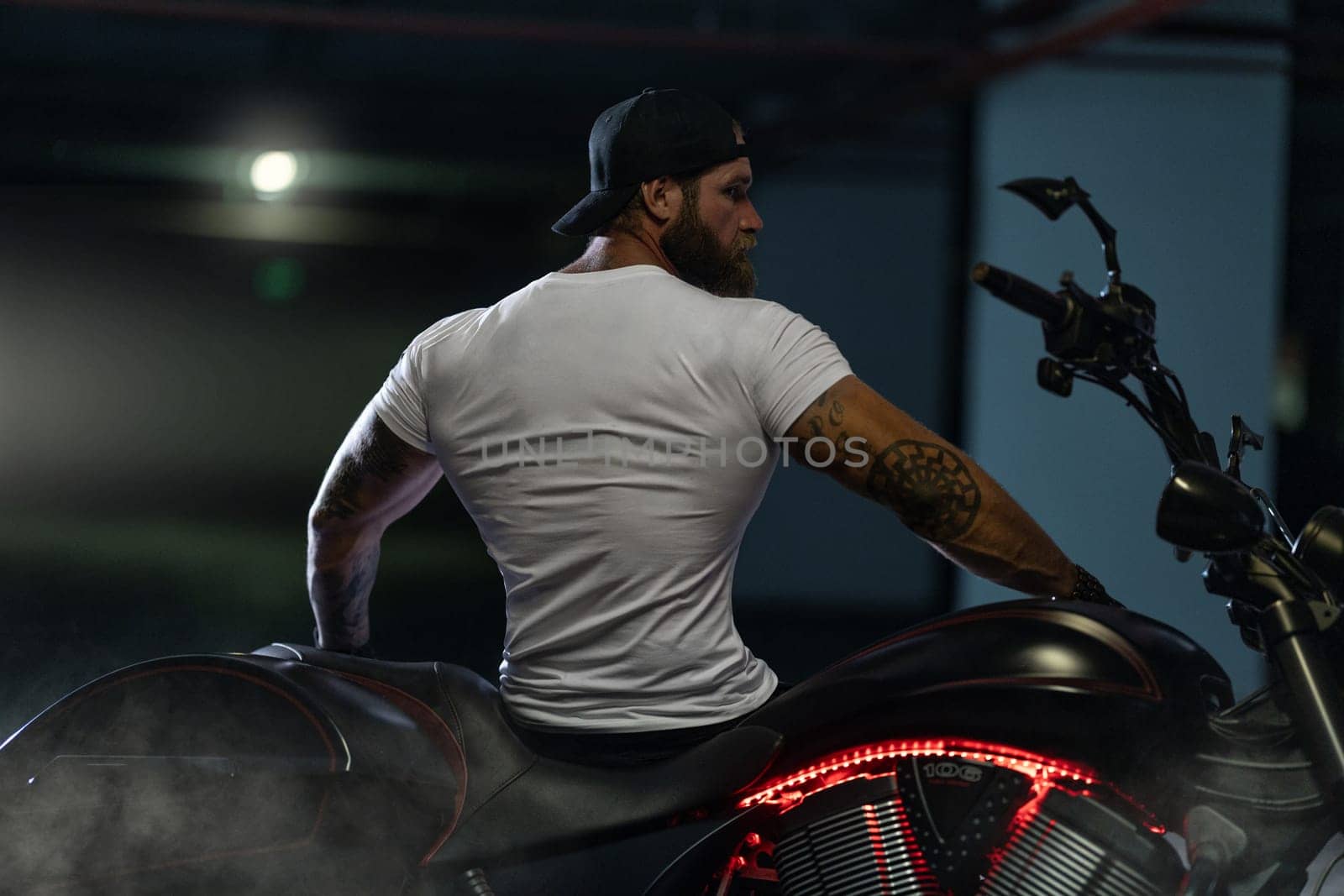 Sexy handsome attractive sporty muscular fitness model breaded biker with brutal beautiful motorcycle in the parking posing with a motorbike with his back to the frame